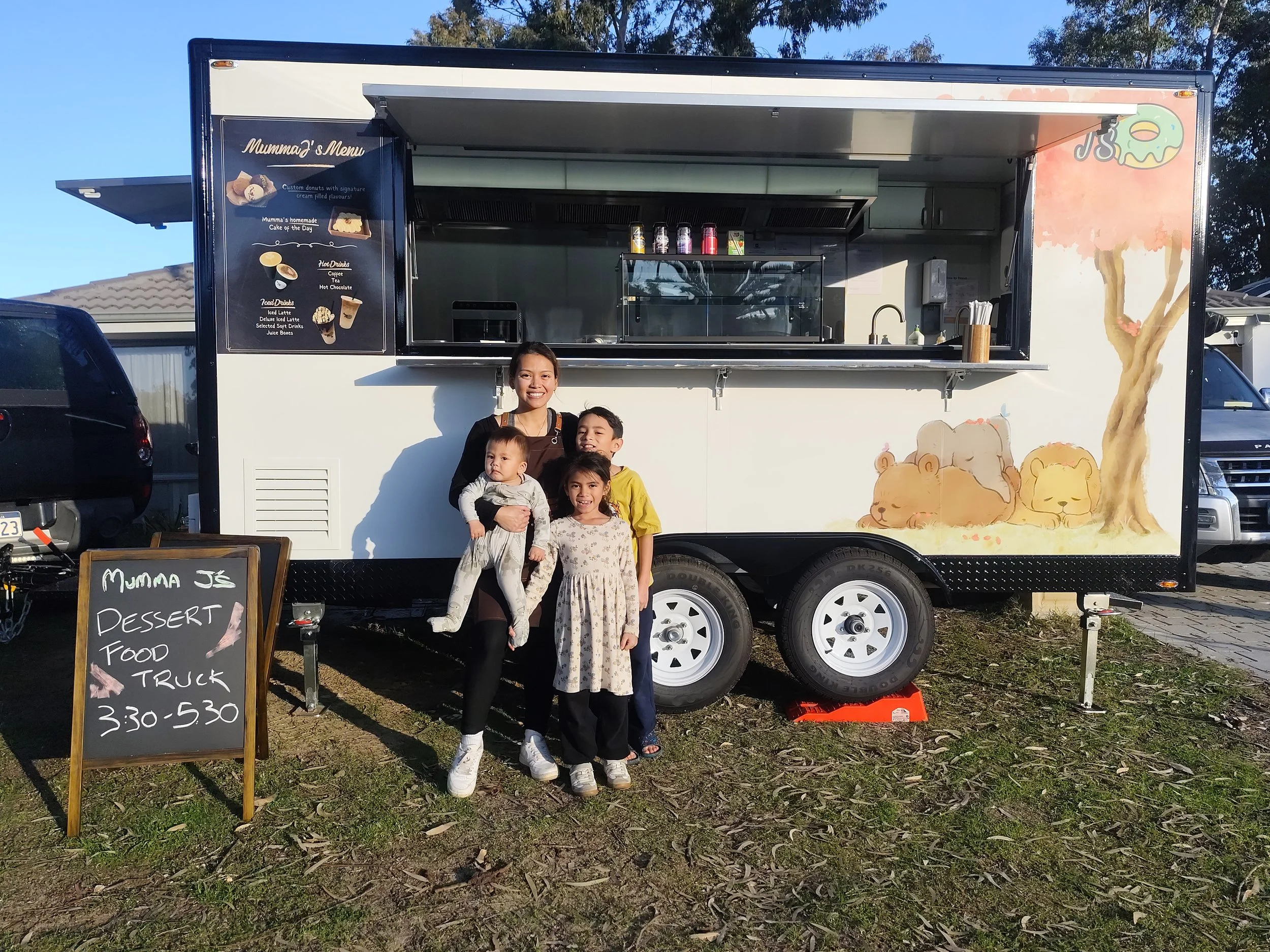 Family standing outside a dessert food truck decorated with cartoon animals and a tree, with a chalkboard sign indicating dessert hours, on a sunny day.