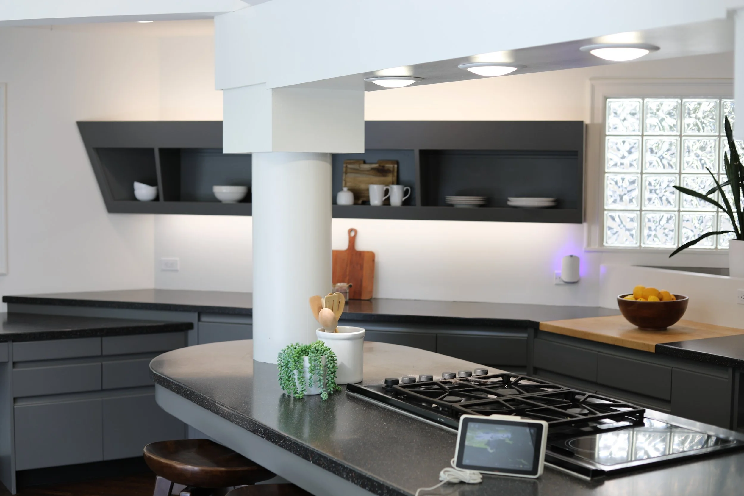 Modern kitchen with dark gray cabinets, open shelves, and a kitchen island with a gas stove, small green plant, and decorative items. There is a wooden bowl with yellow fruits near the window, and a small electronic device on the countertop.