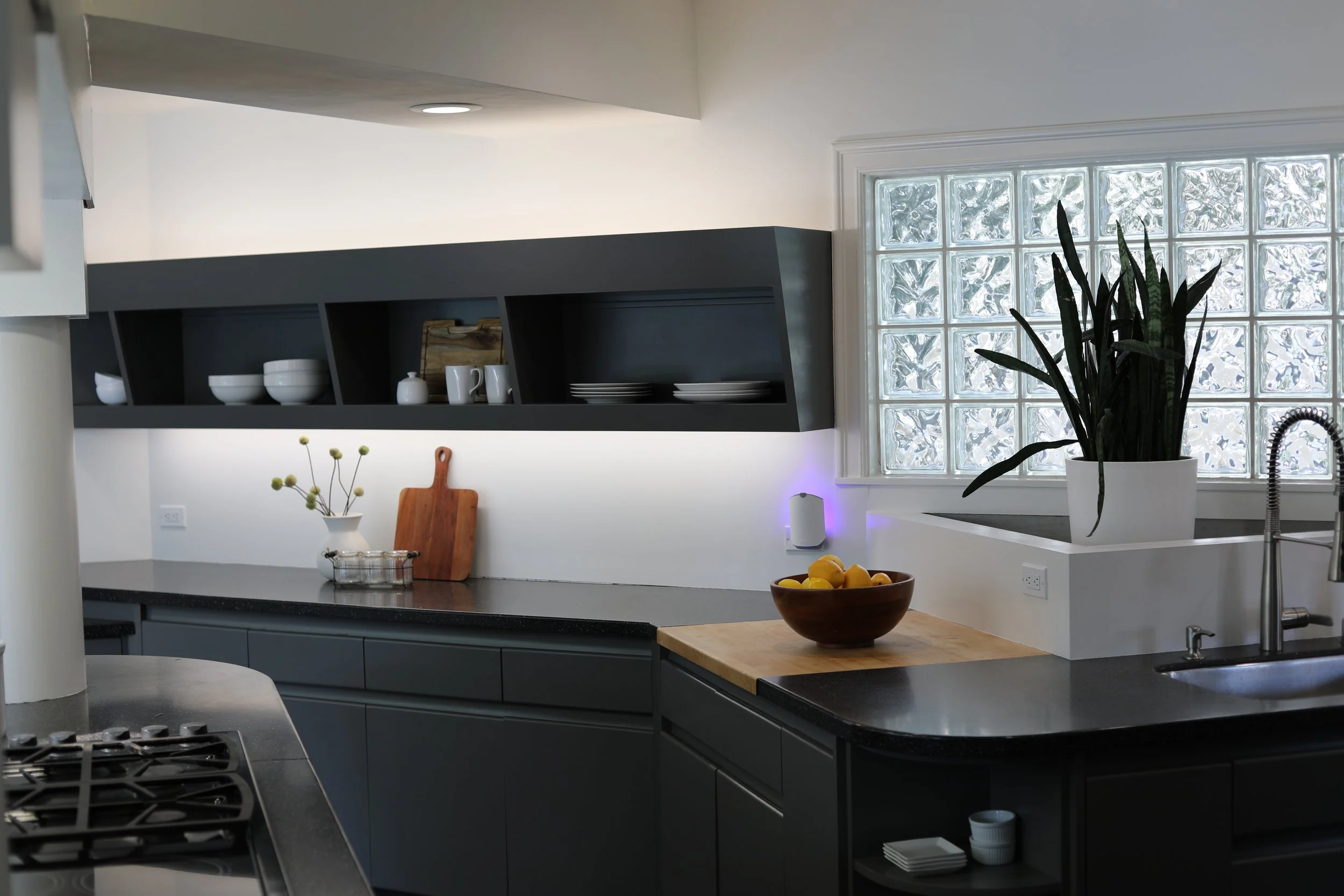 Modern kitchen with black cabinets, open shelving, a window with glass blocks, a large potted plant, and a bowl of lemons.