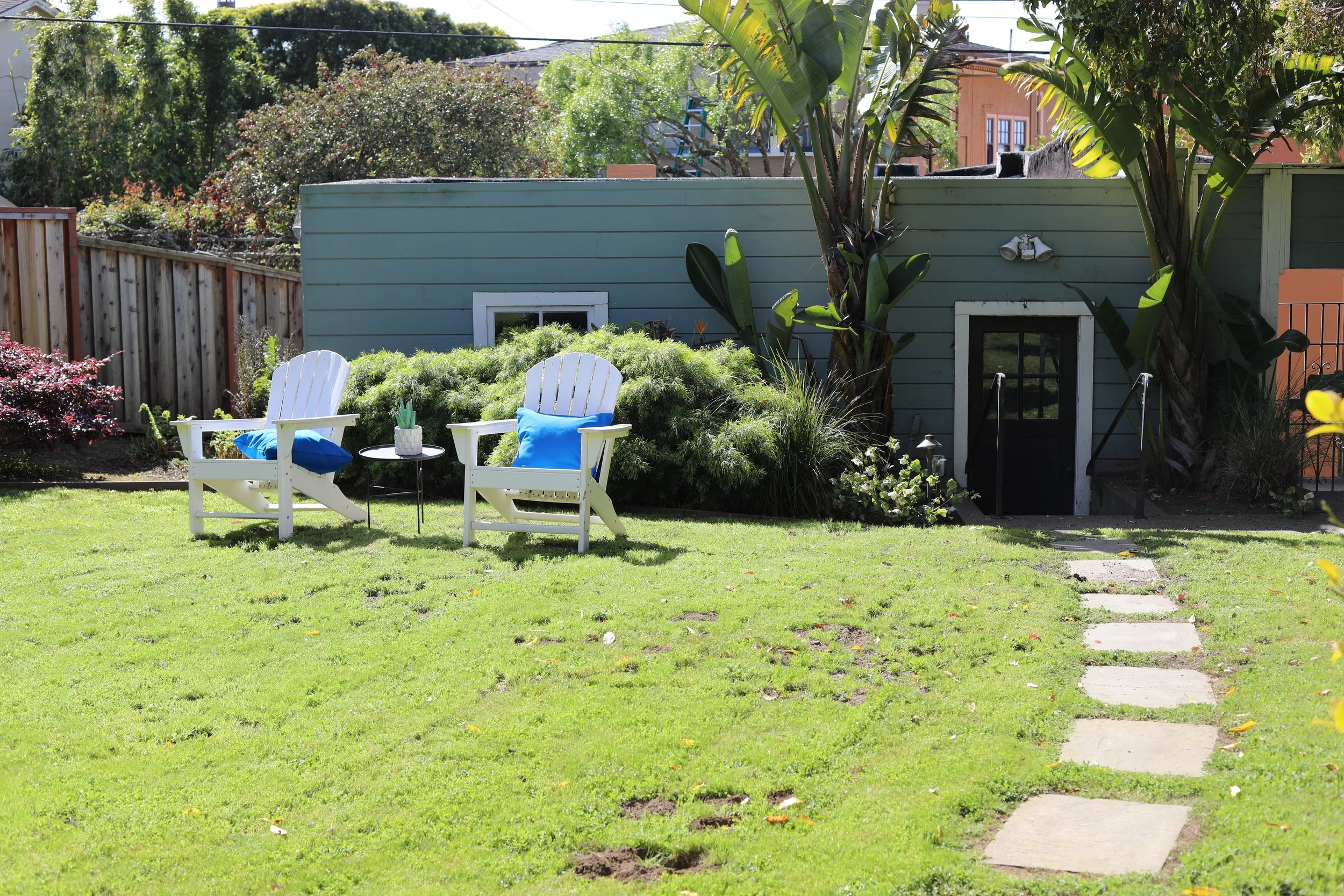 A backyard with green grass, stepping stones leading to a small shed, and two white Adirondack chairs with blue cushions and a small black table between them. There are green plants, bushes, and trees surrounding the area, with a wooden fence on one 