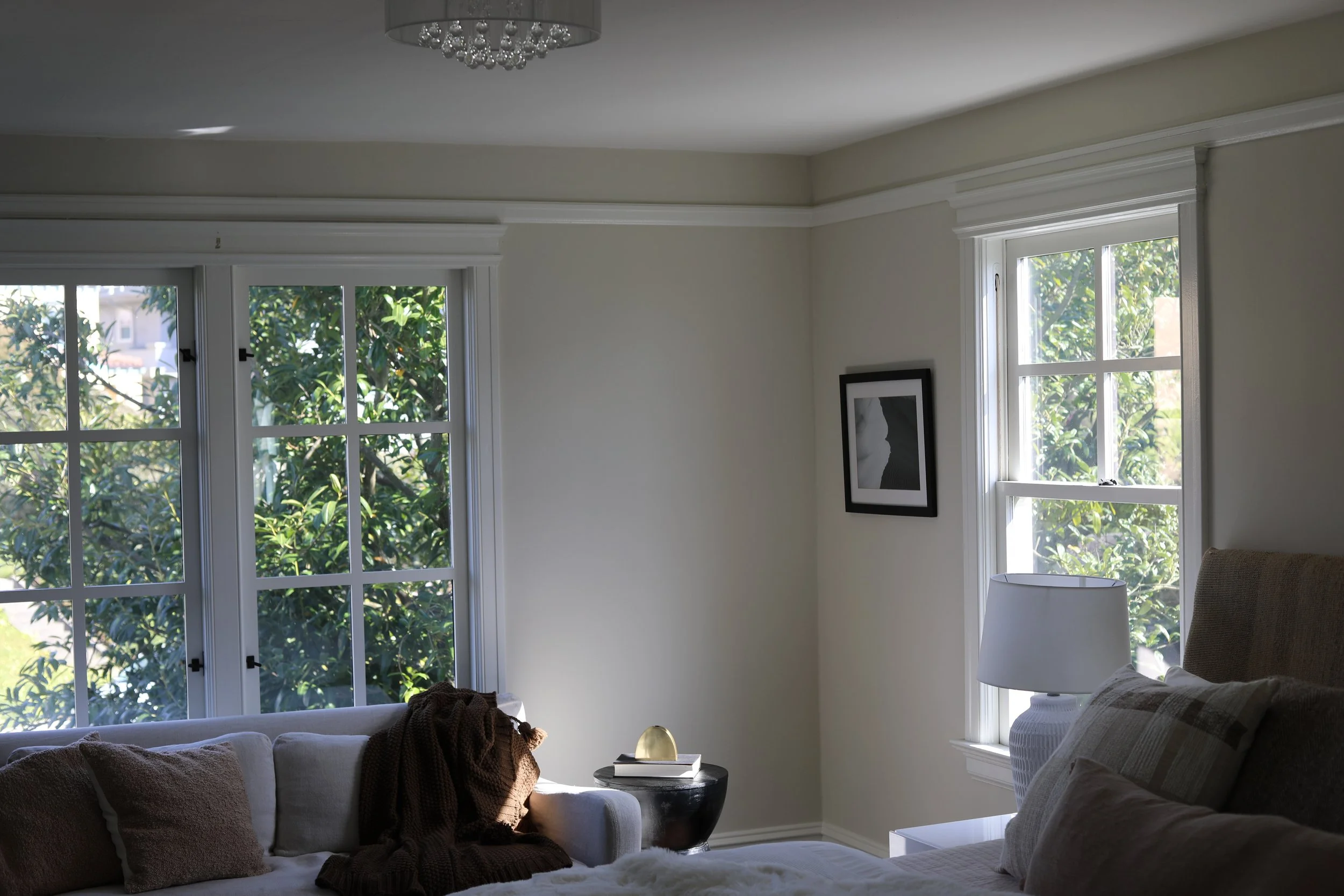 Bright living room with large windows showing greenery outside, a white sofa with brown blanket and pillows, a black side table with a decorative item, a white bedside table with a lamp, and a picture frame on the wall.