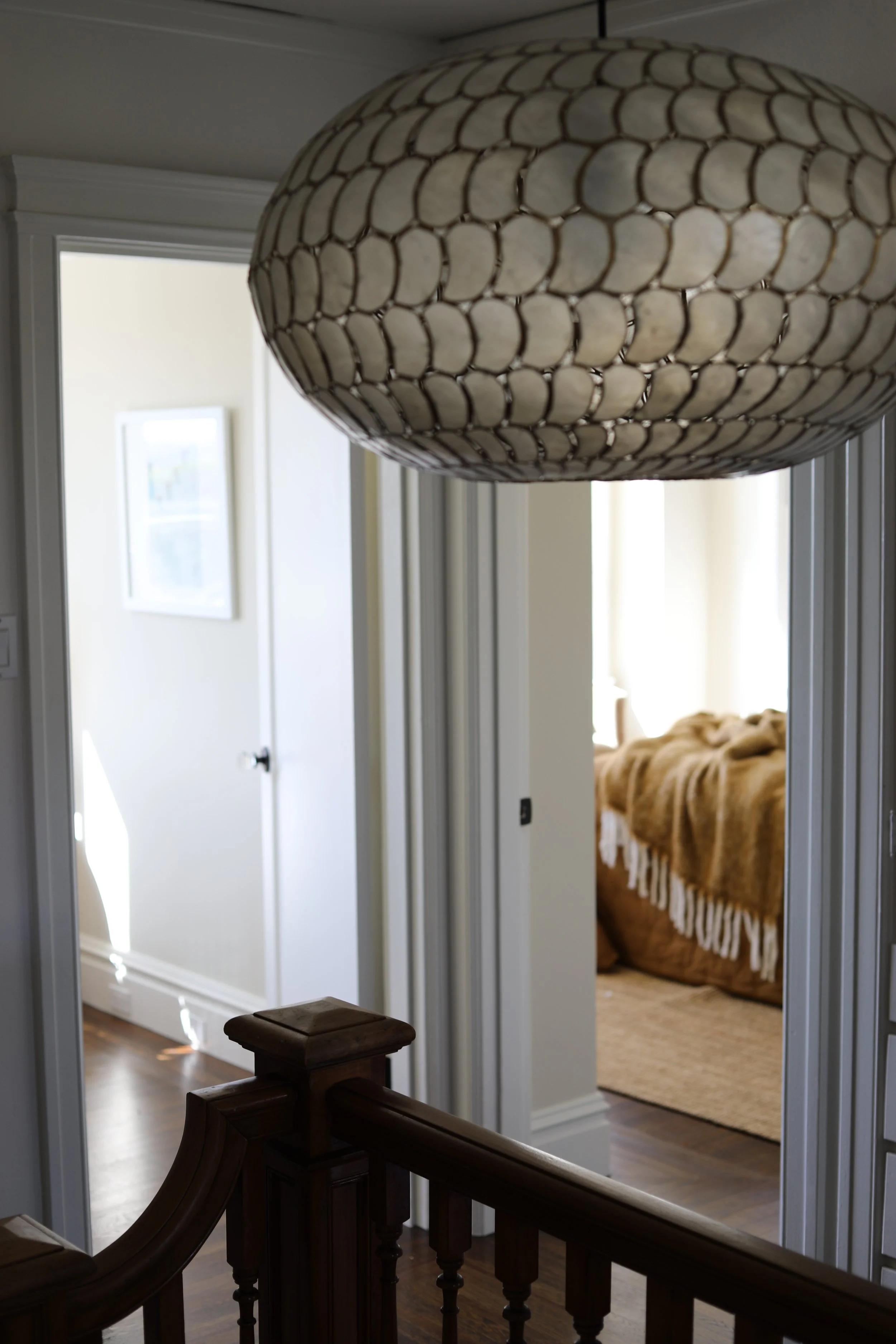 A view from the stairwell showing a hanging ceiling lamp with a shell-like pattern, a hallway with two doors, and a bedroom with a bed covered in a brown blanket.