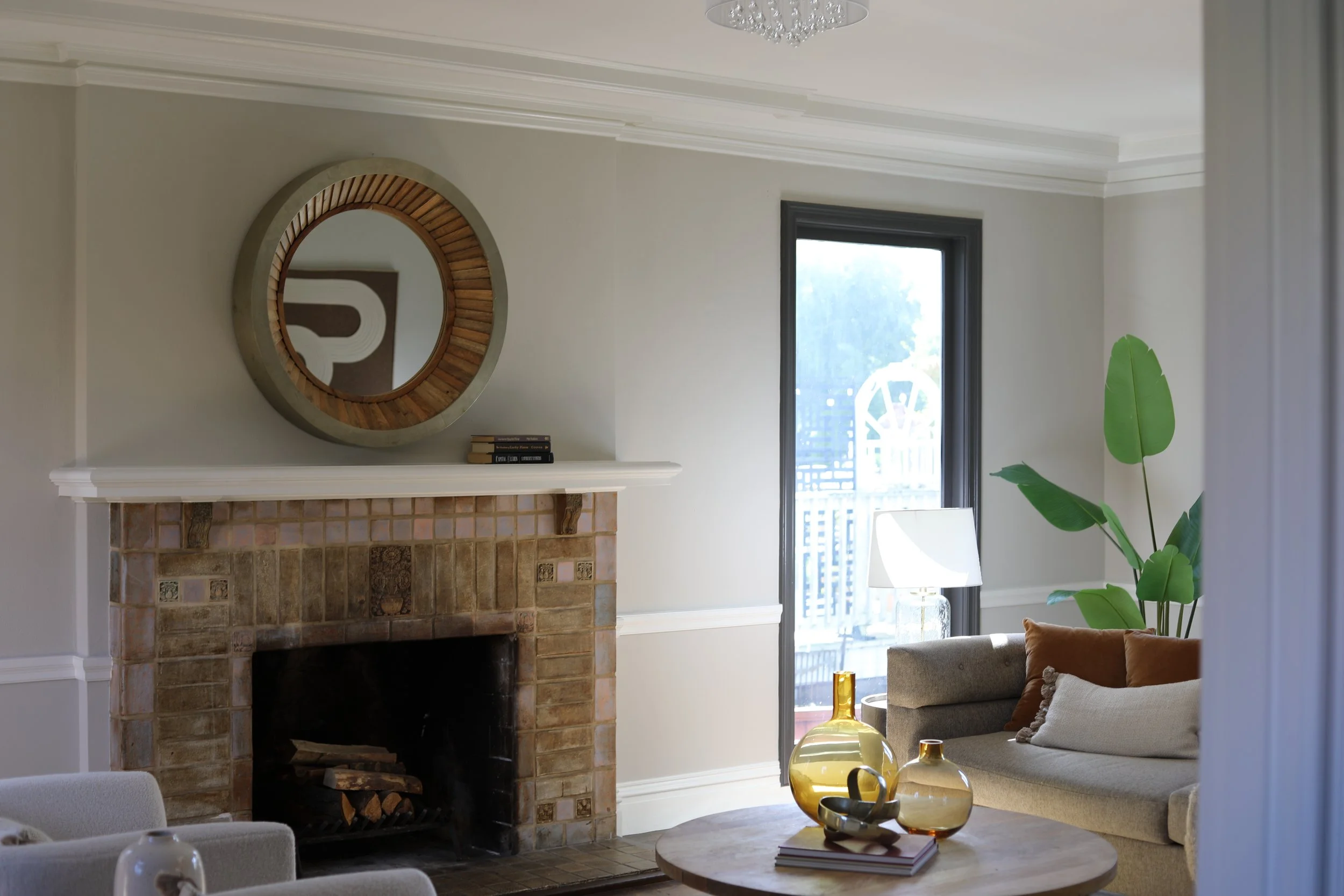 Living room with fireplace, round mirror above mantle, large green plant, window with sunlight, beige sofa with cushions, side table with yellow vases, and a white side chair.
