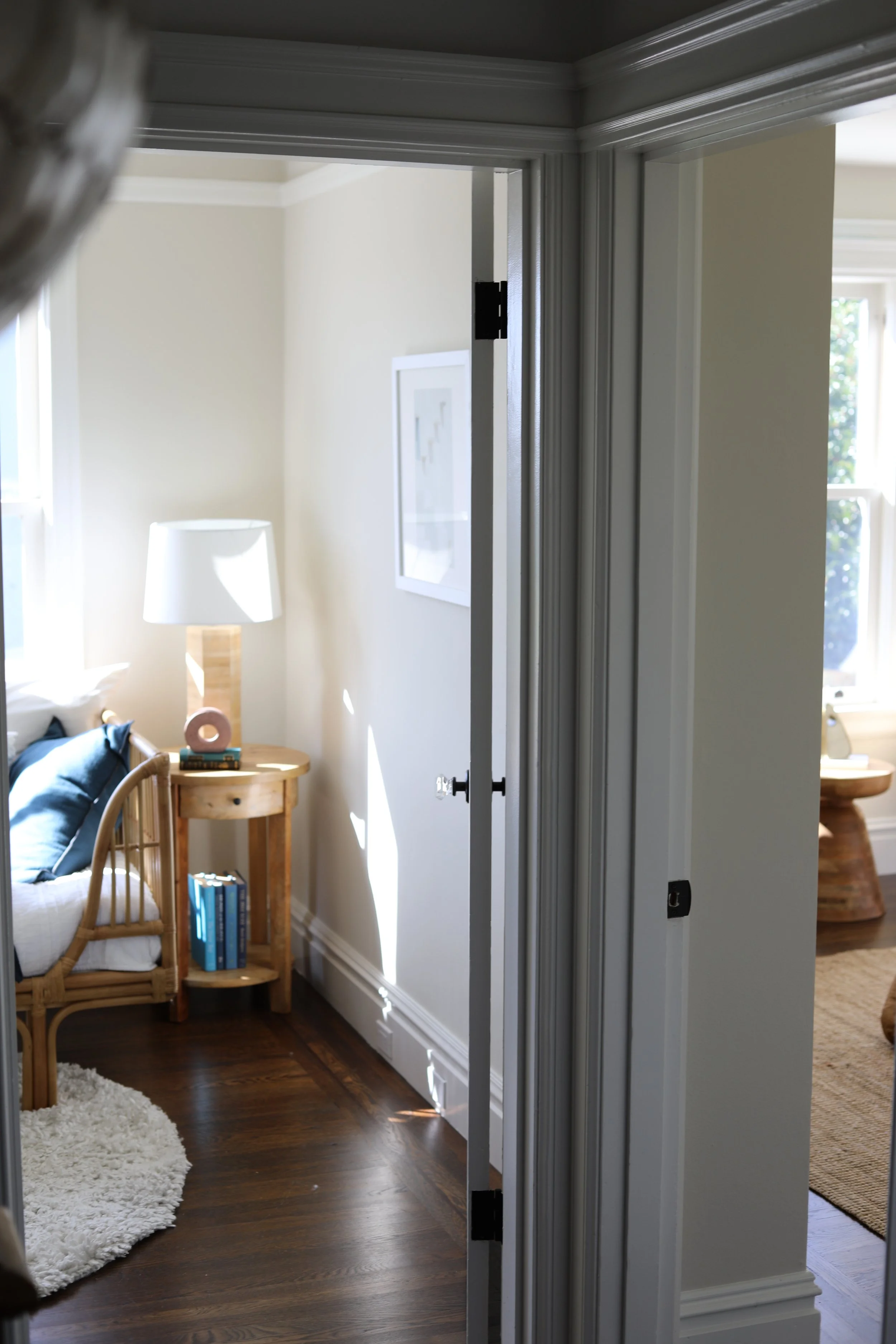 Interior view of a room with a door frame, showing a corner with a wooden side table, a white lamp, a framed picture on the wall, and a bed with pillows, sunlight coming through the windows, and a wood floor.