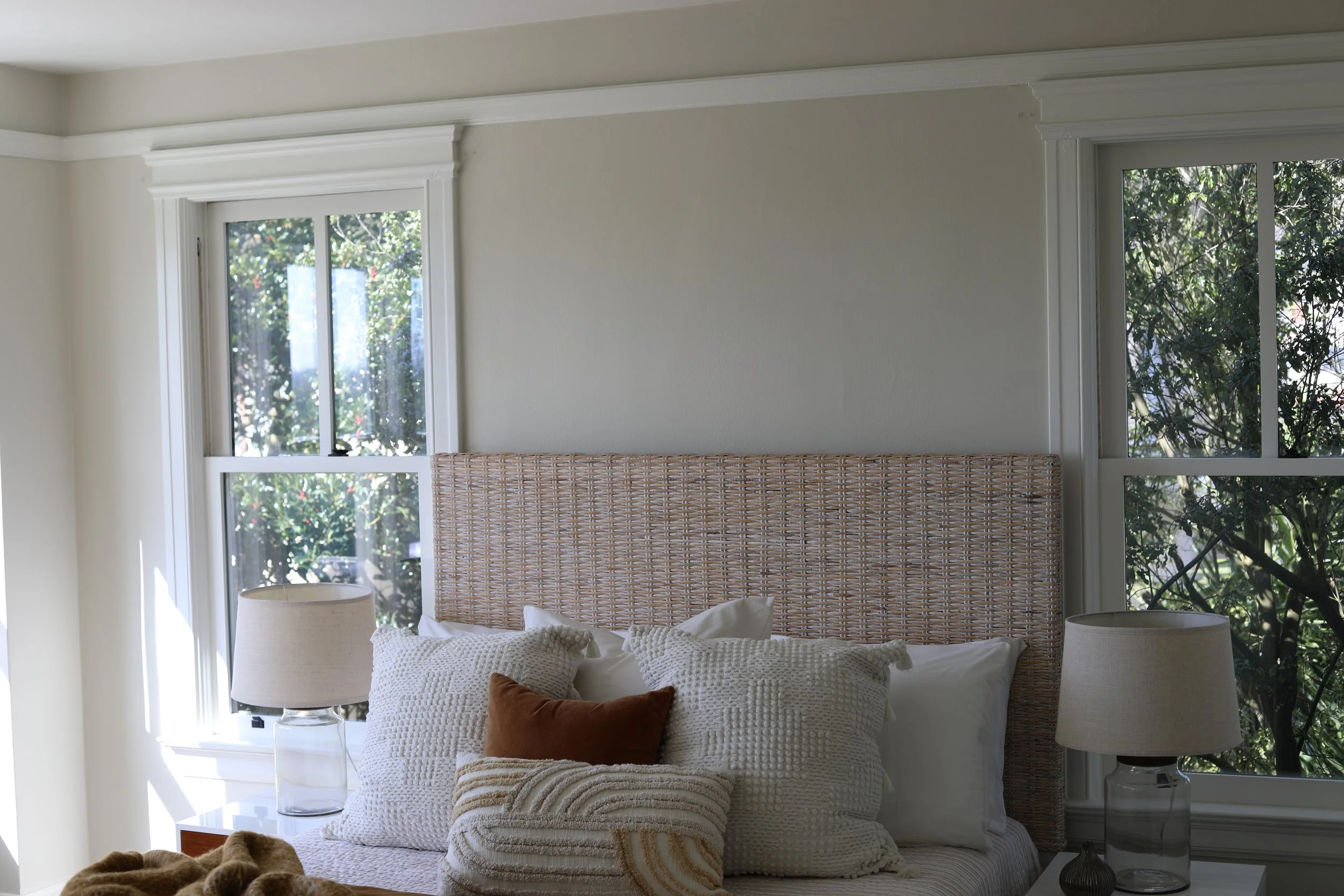A bedroom with two windows, a tan woven headboard, white walls, and two glass table lamps with white shades on bedside tables. There are multiple pillows on the bed, including a brown one and textured white pillows, and a blanket at the foot of the b