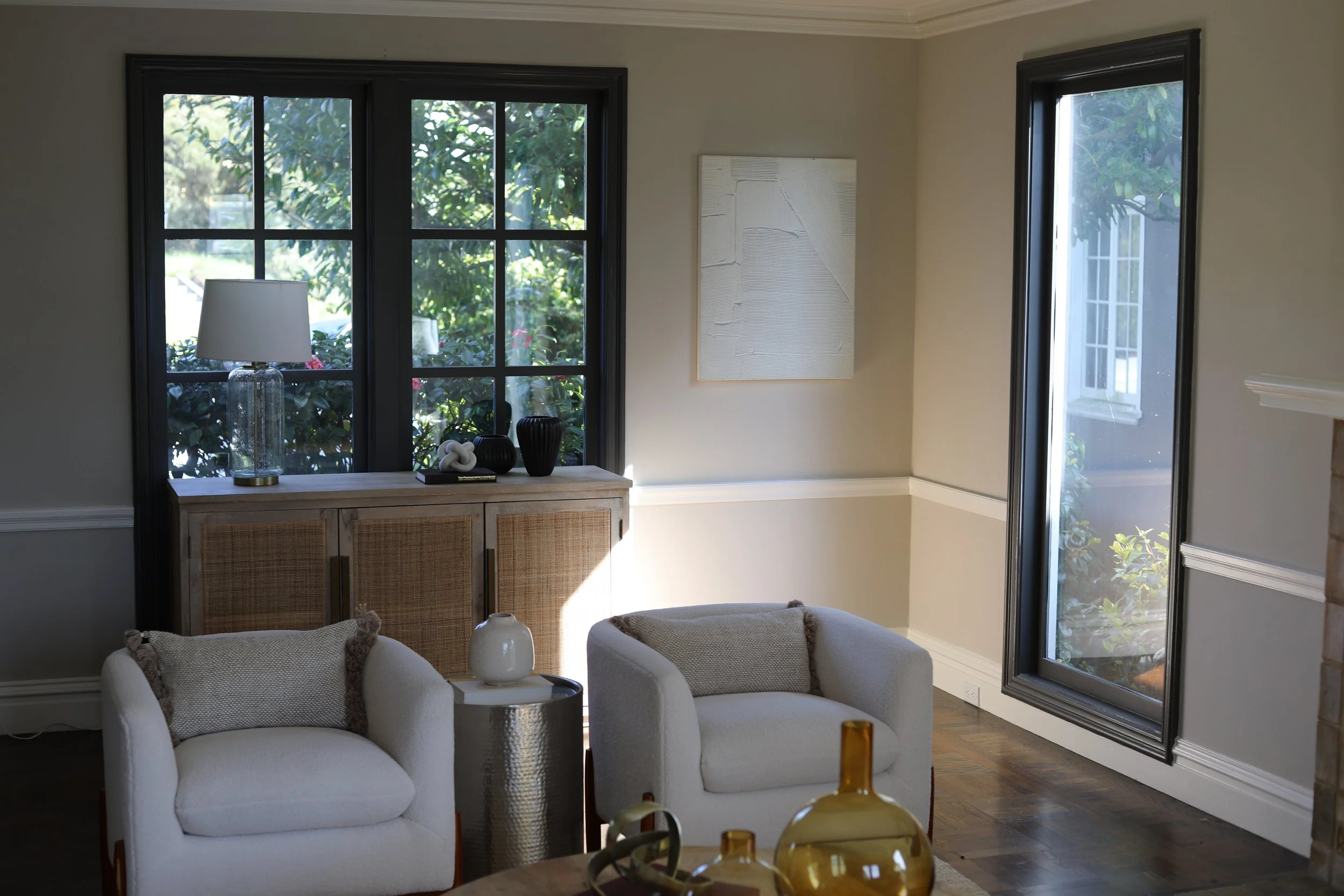 Living room with two white armchairs, a small round side table, a wooden cabinet with decorative items, large window with black frame, and a glass sliding door, with sunlight streaming in.