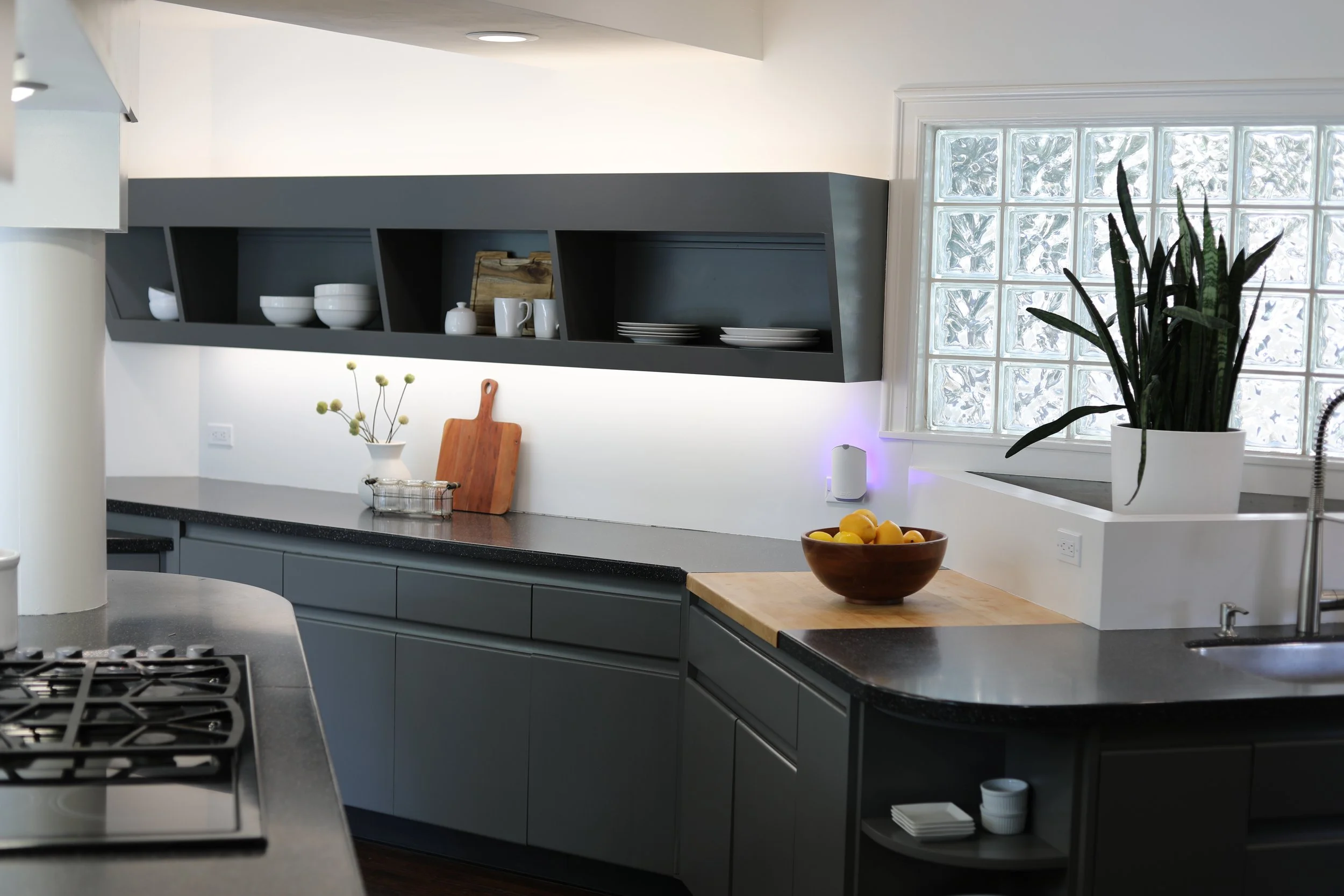 Modern kitchen with gray cabinets, open shelving, a wooden cutting board, a bowl of lemons, and a large plant near a glass block window.