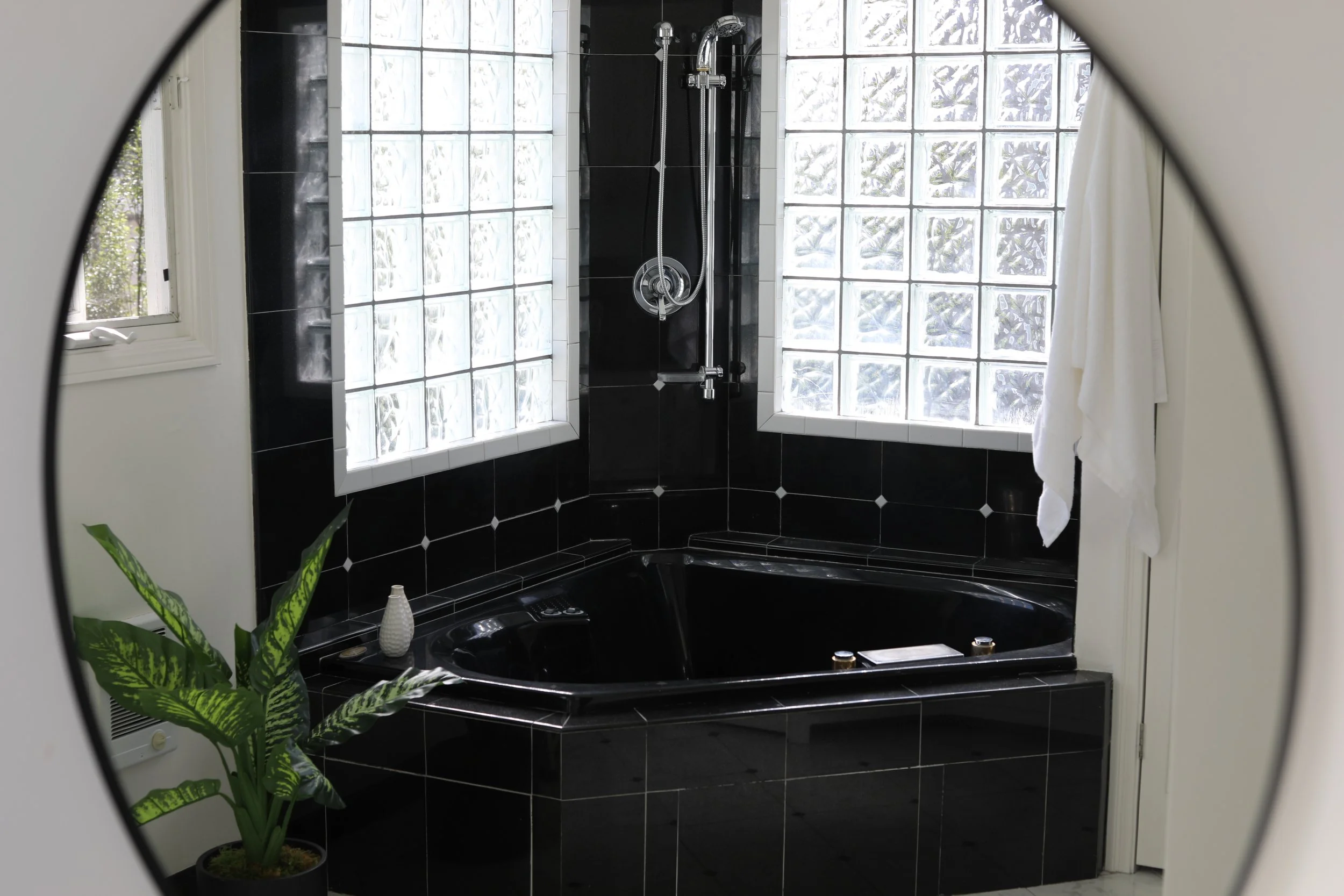 A black bathtub in a bathroom with black tiled walls and glass block windows, a white towel hanging on the wall, and a green potted plant next to the bathtub.