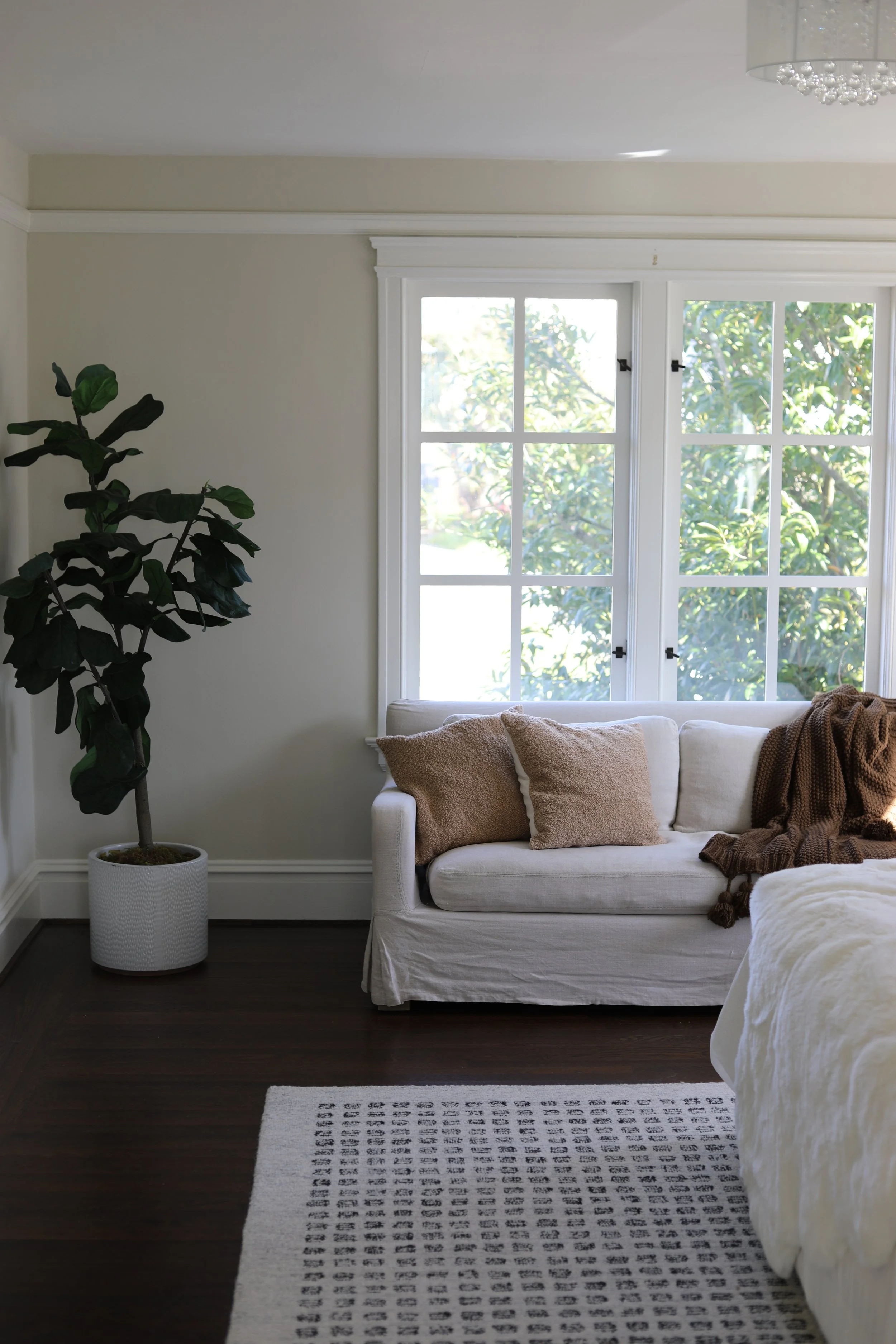 Living room with a white sofa, beige cushions, a brown throw blanket, a large window with greenery outside, a potted plant, a patterned rug, and part of a bed in the foreground.