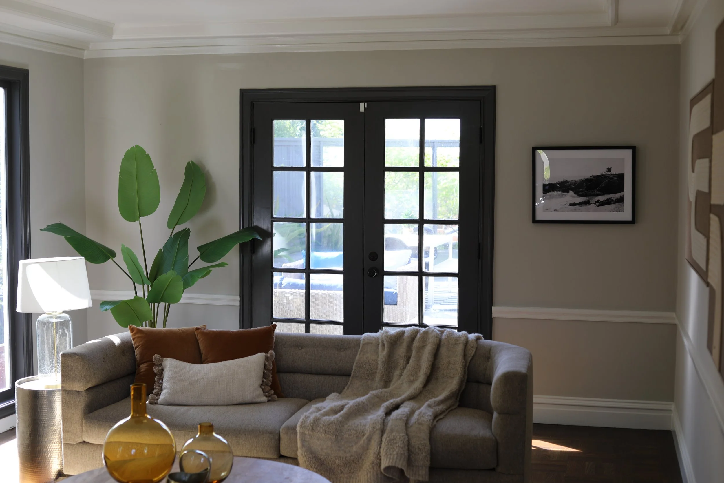 Living room with a beige sofa, decorative pillows, a throw blanket, a tall green houseplant, a glass table lamp, and round decorative vases on a coffee table, with black double glass doors and a framed black and white photograph on the wall.