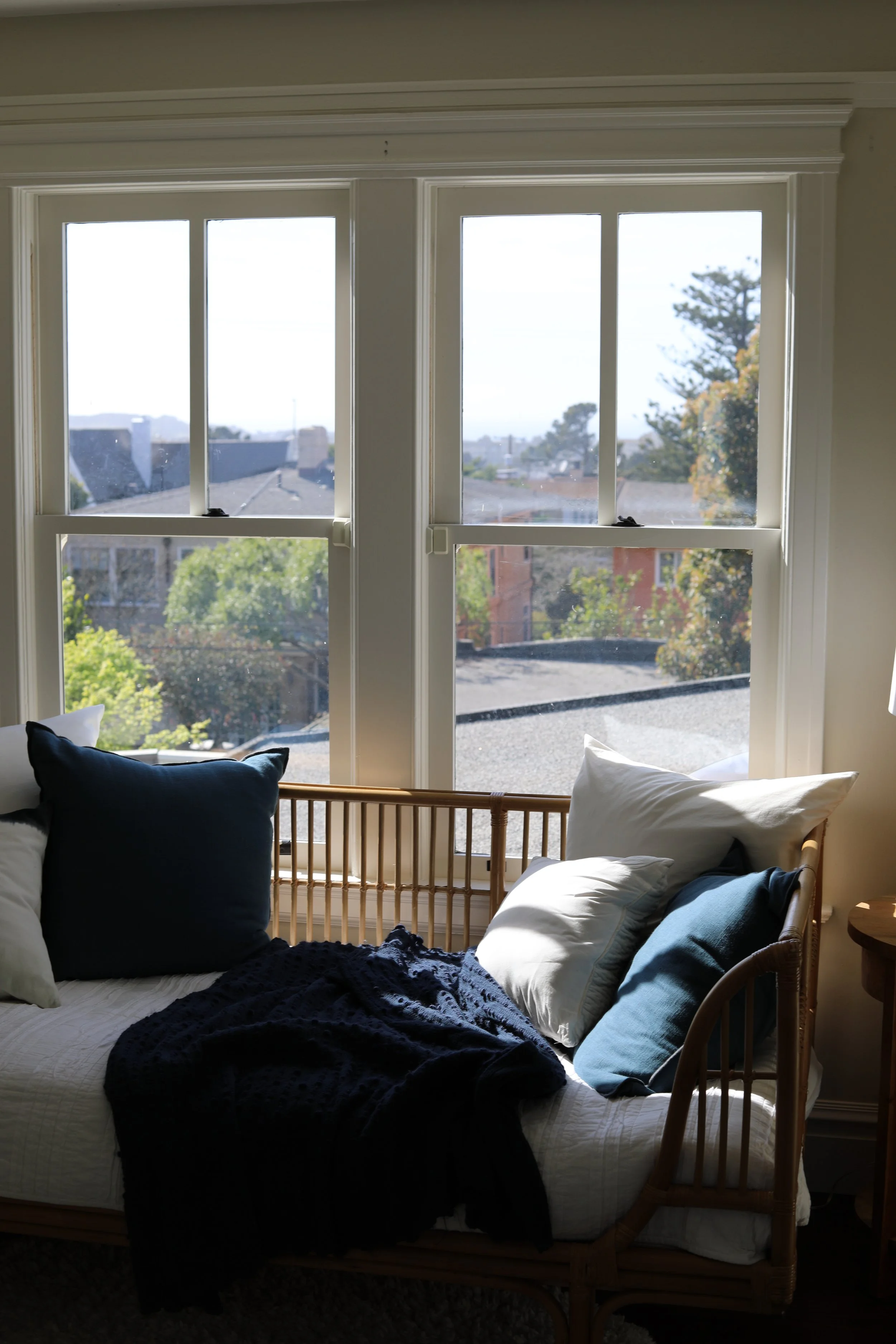 A cozy window seat with pillows and a blanket, overlooking a suburban neighborhood with trees and houses.