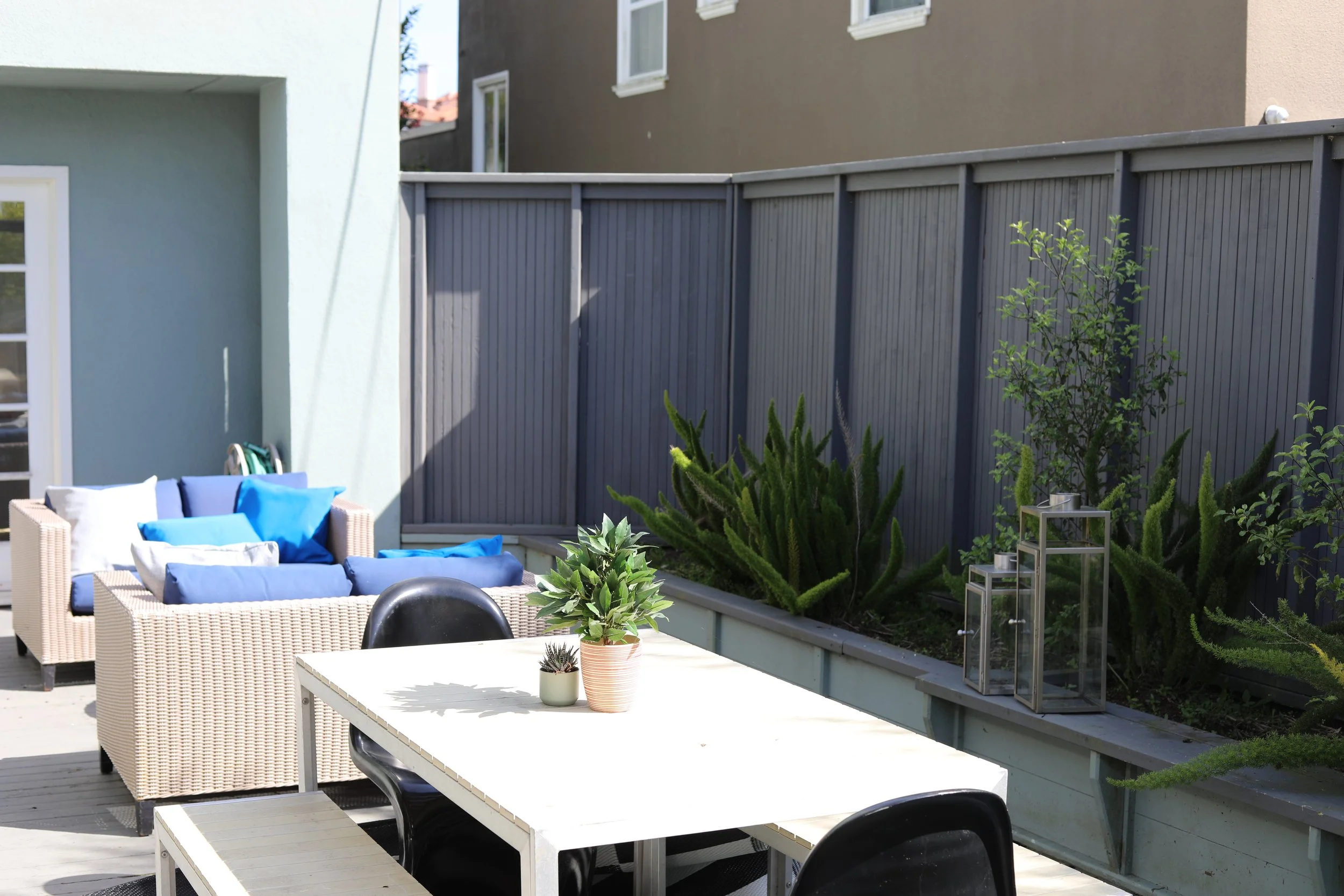 Outdoor patio area with a white table, black chairs, and a white wicker sofa with blue cushions, surrounded by green plants and a gray privacy fence.