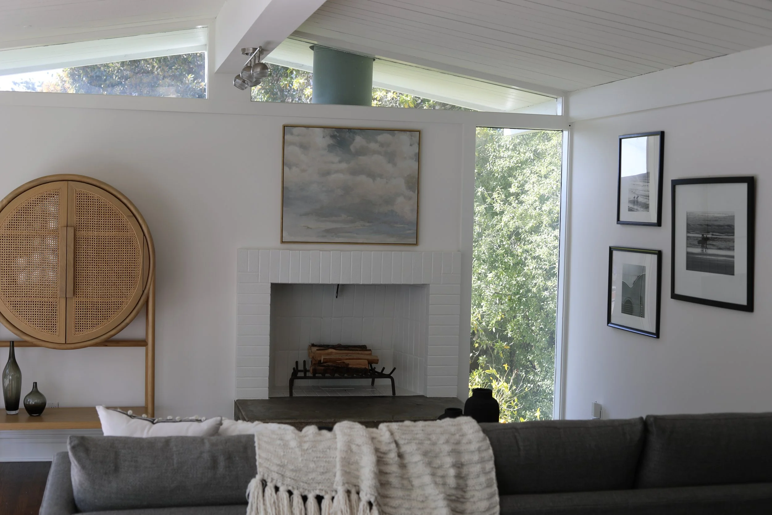 Living room interior with a gray sofa, a white throw blanket, and decorative vases. There is a white brick fireplace with a stack of firewood inside. Artwork hangs above the fireplace and on the adjacent white wall, with large windows showing greener