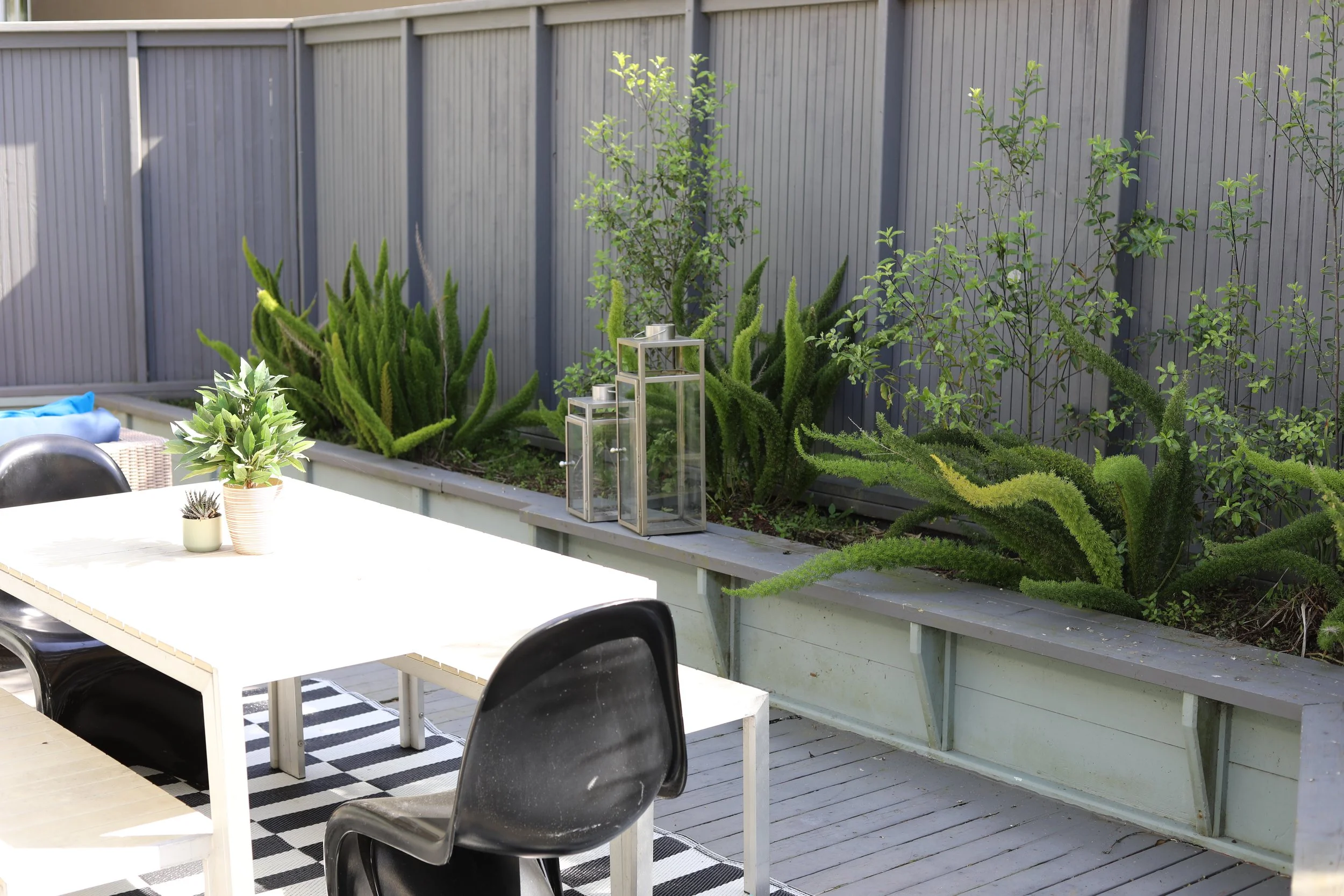 Outdoor patio with a white dining table, black chairs, potted plants, and decorative lanterns, with a raised garden bed filled with green succulents and small trees against a gray wooden fence.
