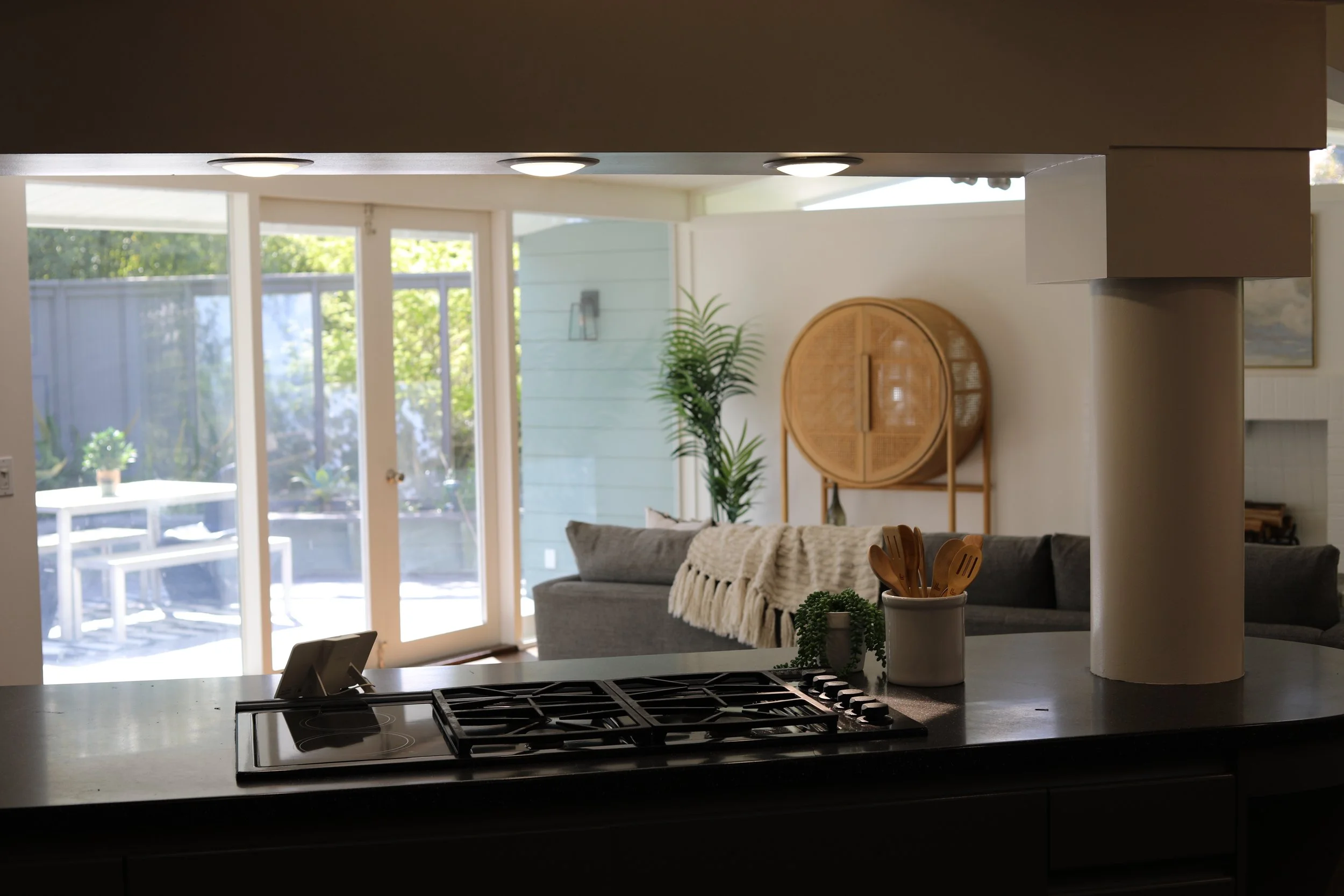View of a modern open-concept kitchen and living room with sliding glass door leading outside, featuring a potted plant, wooden decor piece, and neutral-colored furniture.