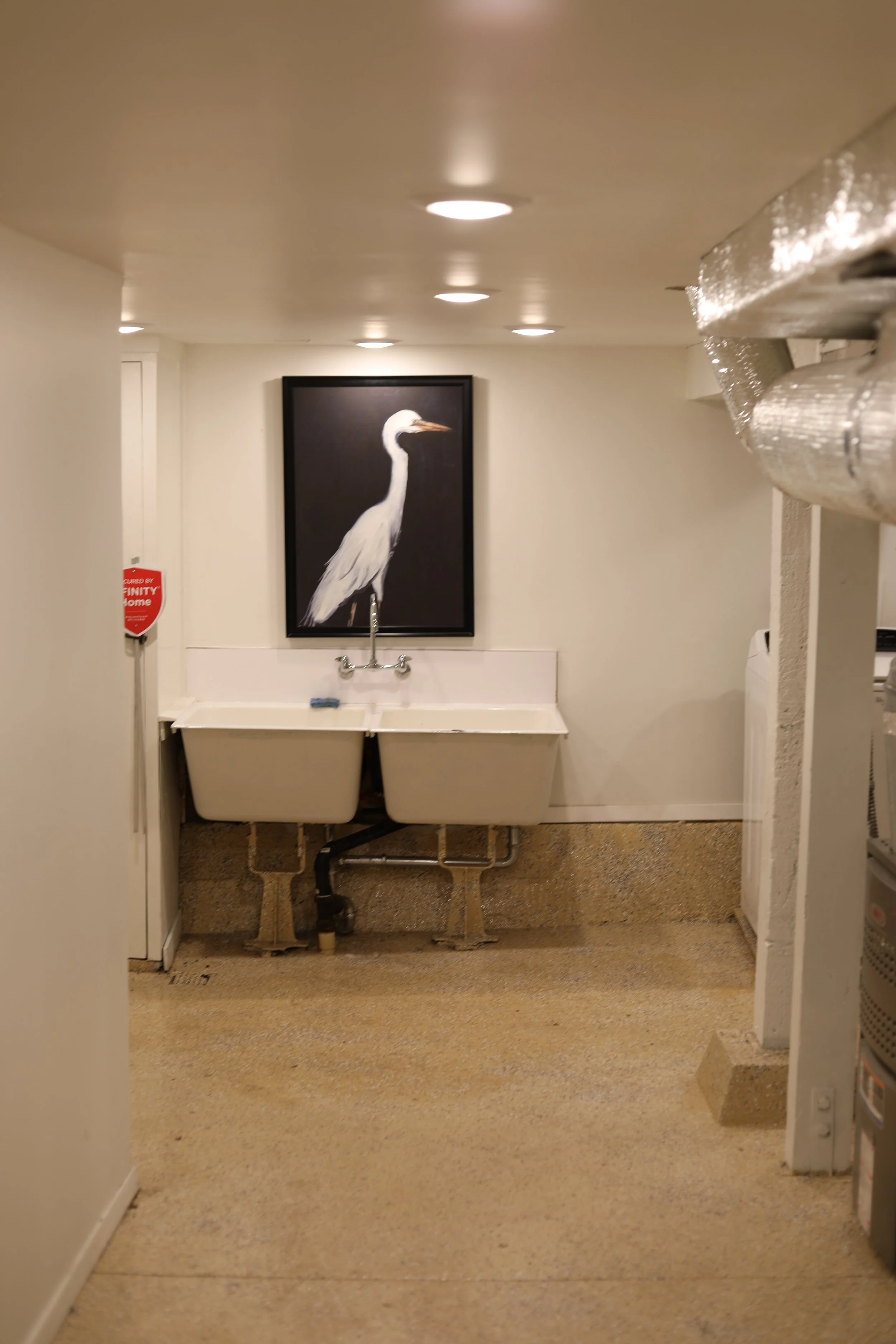 A laundry room with a framed picture of a heron on the wall, a utility sink with a faucet, and various pipes and ducts.