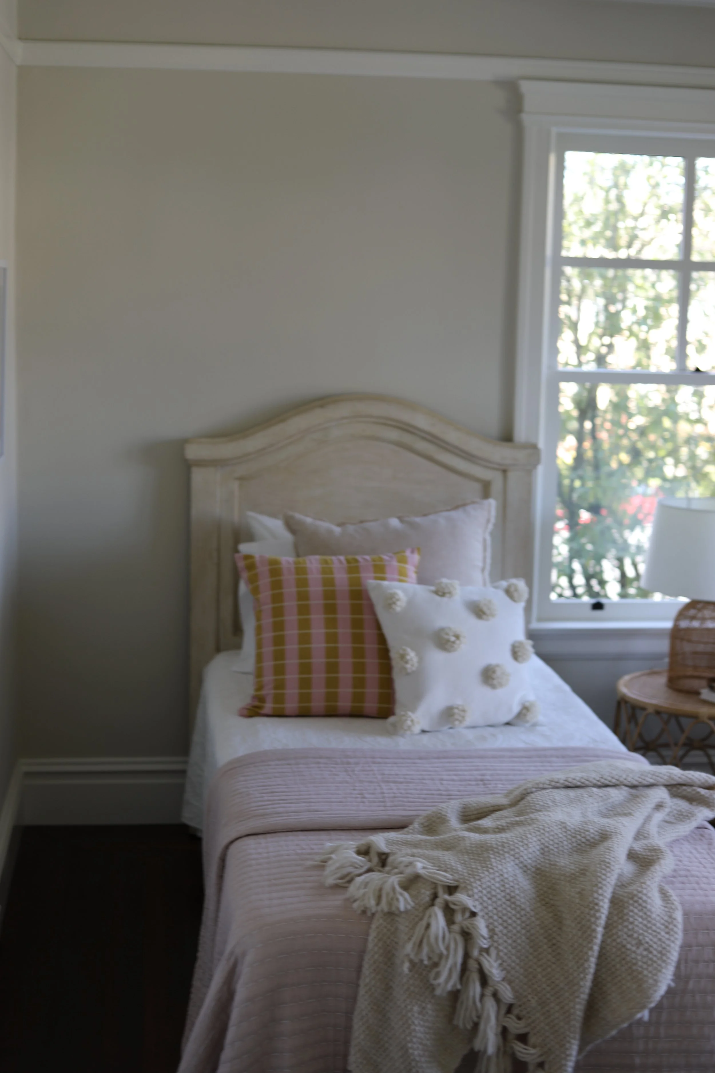 A cozy bedroom with a beige wooden headboard, decorated with pillows including one with pink and yellow plaid, a white pillow with pom-poms, and an additional pillow behind. There is a window with white trim allowing natural light to fill the room, a