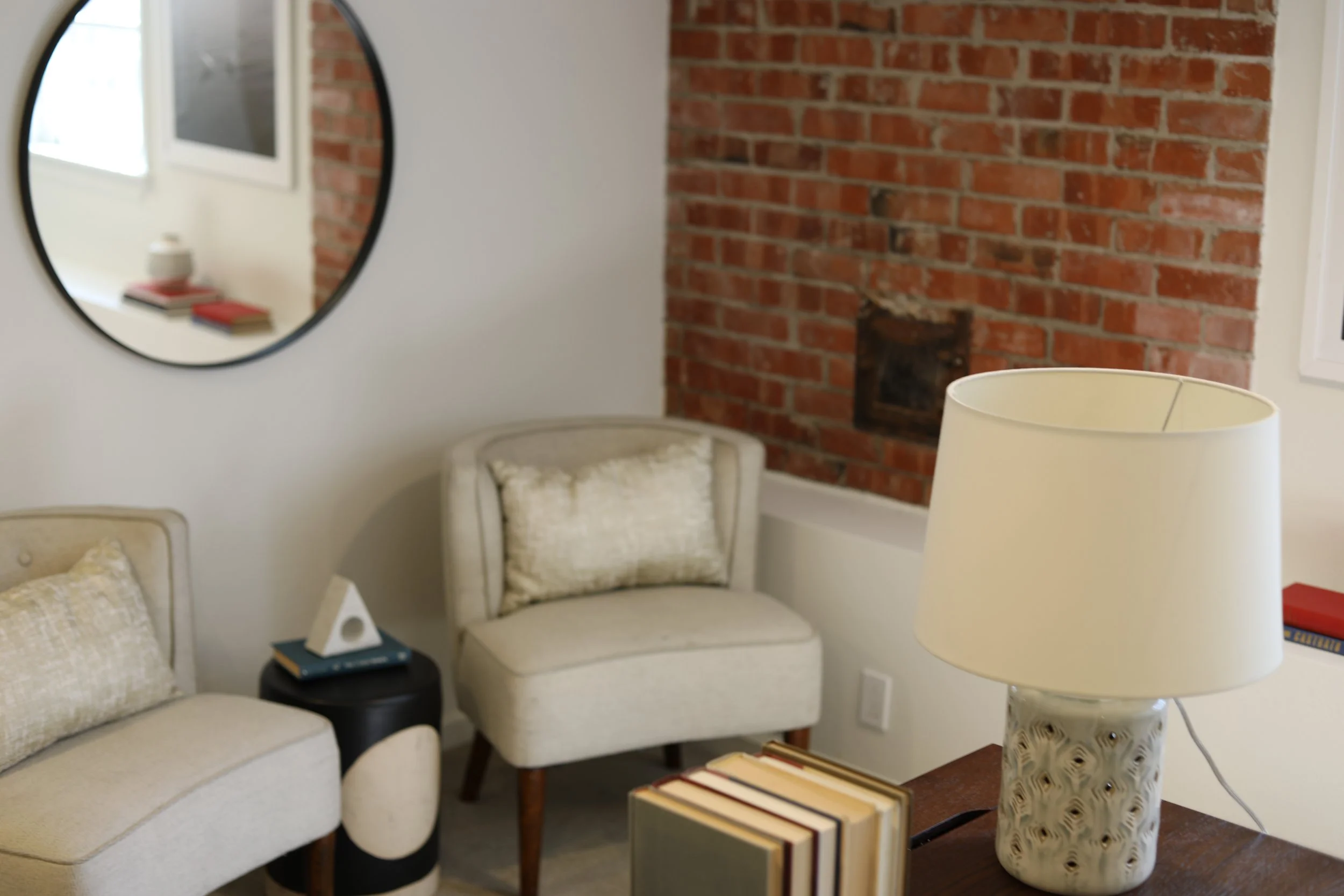 Living room with beige chairs, a black side table, a table lamp with a patterned base, and a brick wall