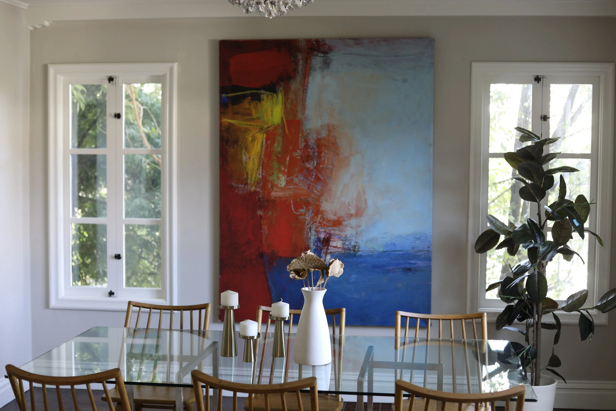 A modern dining room with a glass-top table, wooden chairs, abstract painting with red, yellow, blue, and white colors on the wall, a tall white vase with dried flowers, and a large potted plant next to two windows with white frames, overlooking gree