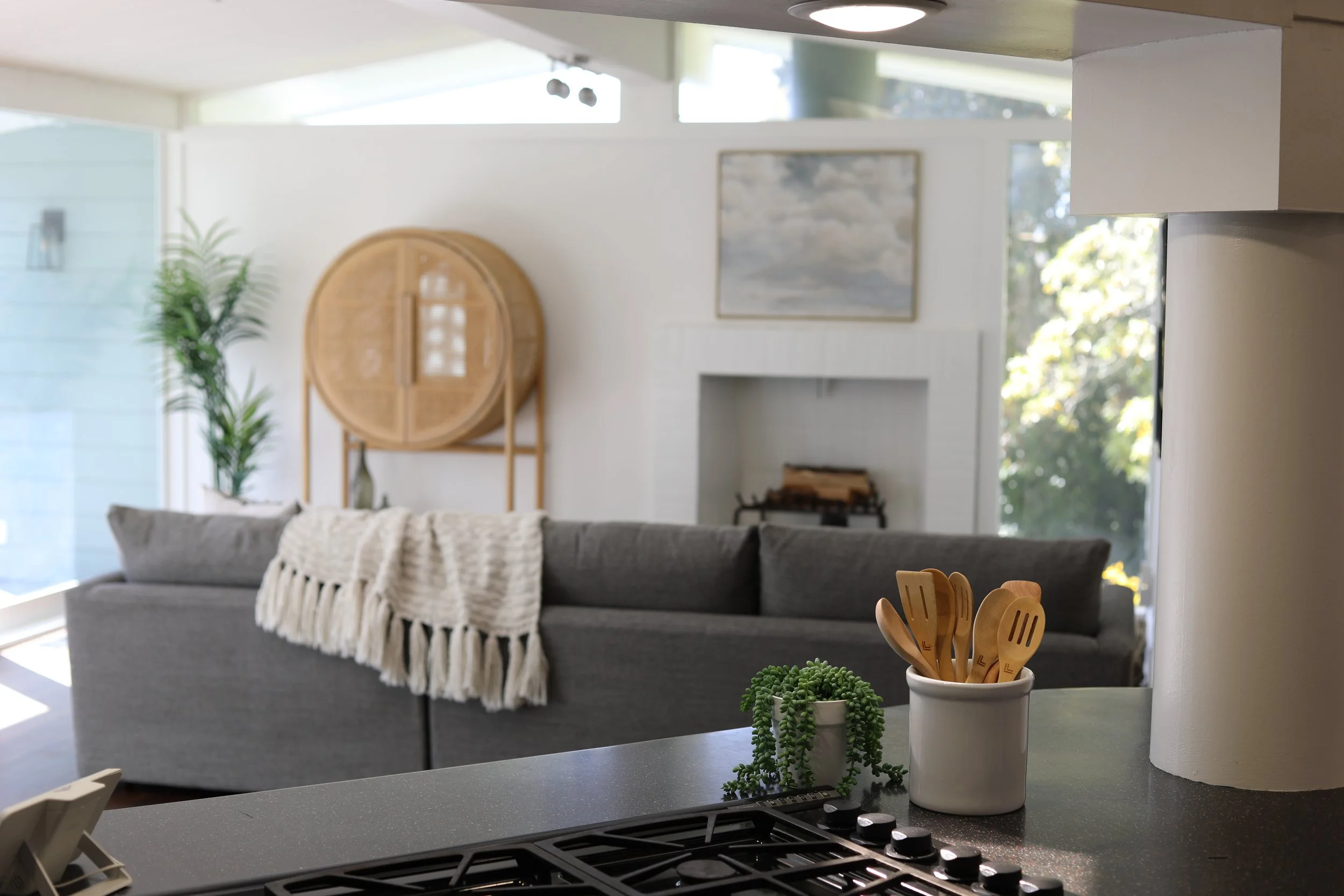 Living room and kitchen view with a gray couch, a blanket, a stove, and a container of wooden utensils, with large windows and indoor plants.