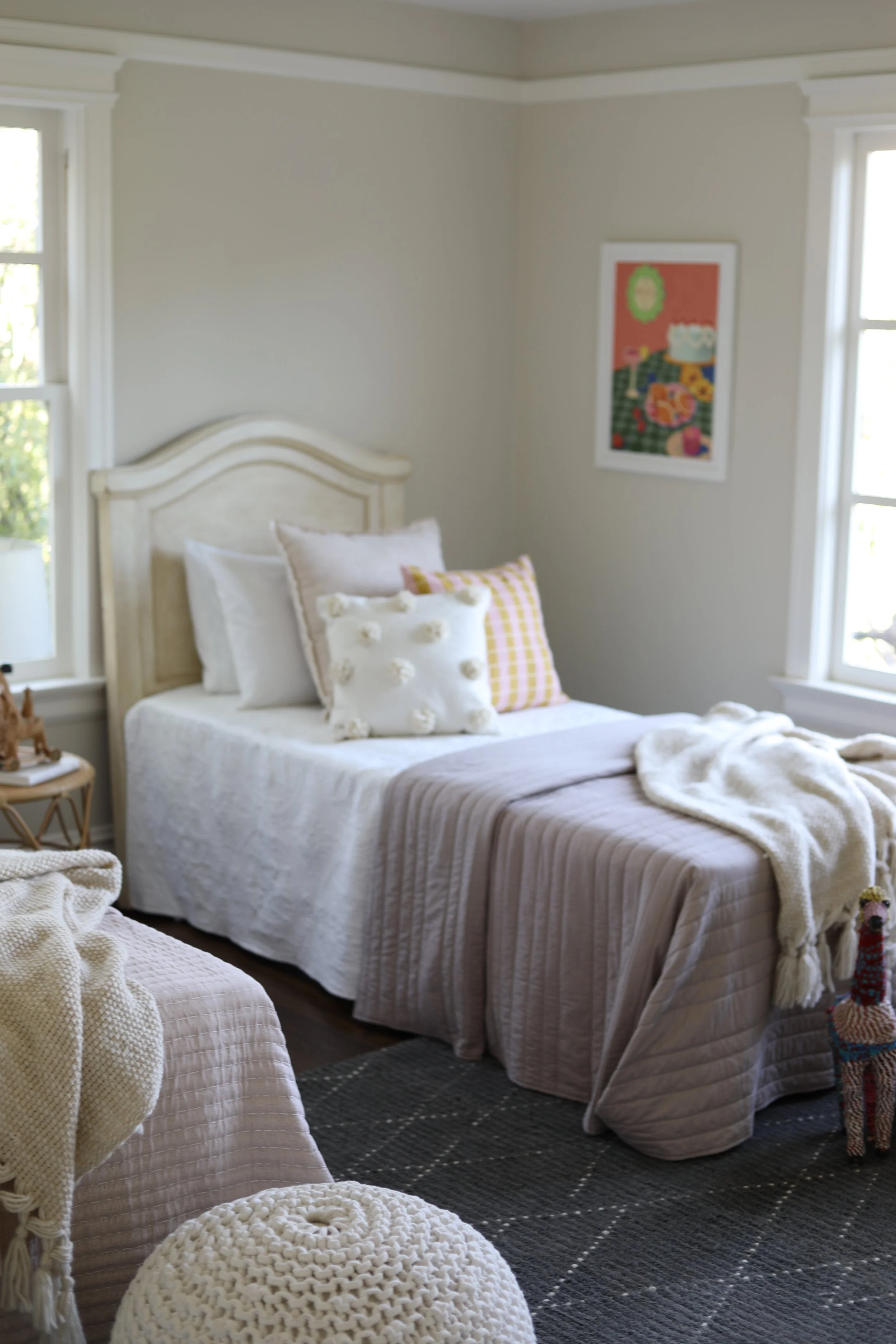A cozy bedroom with a white bed, decorated with pillows and a soft throw blanket. There's a black and white rug on the floor, and a framed colorful artwork hangs on the wall. A knitted pouf is in the foreground and a small stuffed animal sits on the 