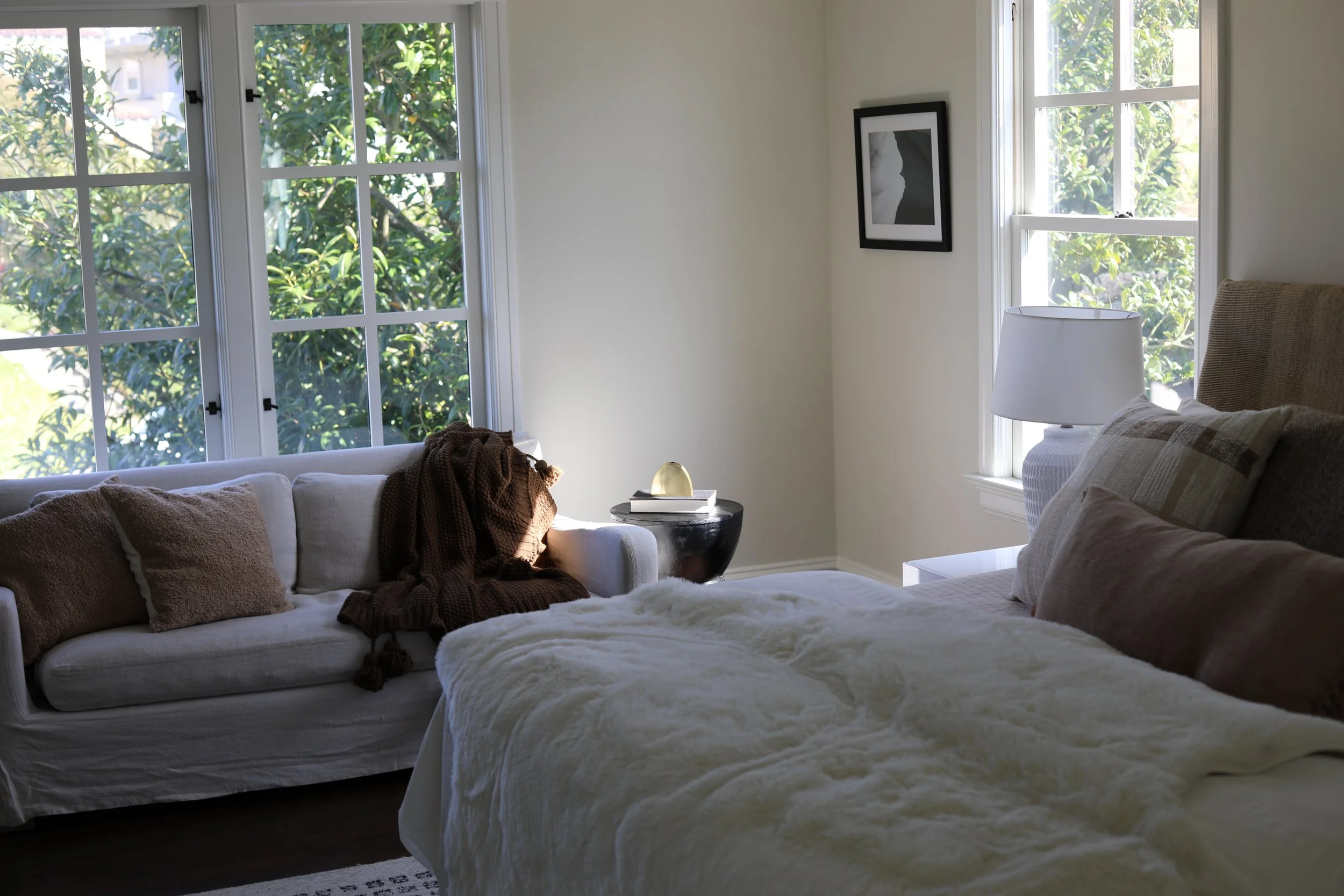 Bright living room with large windows, cream-colored sofa with pillows, a blanket, a small black side table with books and a decorative item, a bed with a white blanket, and a white nightstand with a lamp.
