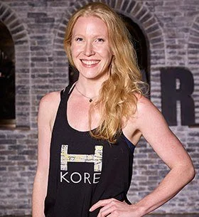 A woman with long red hair smiling at the camera, wearing a black sleeveless top and an apron with the word 'KORE' on it, standing in front of a brick wall.