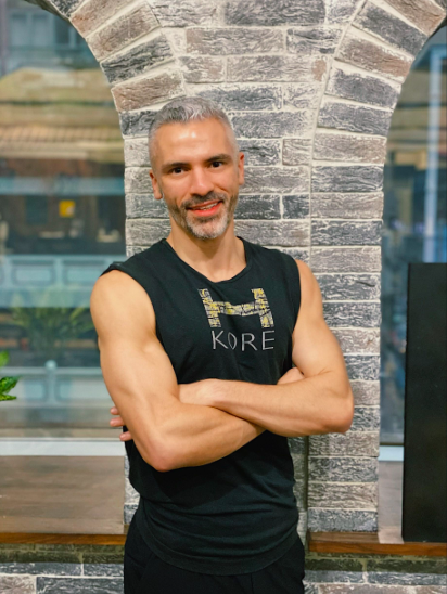 A middle-aged man with short gray hair and a beard stands with arms crossed in front of a brick archway at a modern indoor location.