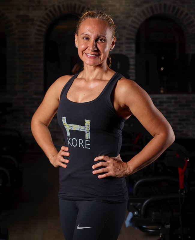 A woman with blonde hair tied back in a ponytail, smiling confidently, standing in a gym with her hands on her hips, wearing a black sleeveless athletic top with a graphic and the word "KORE" on it, and black Nike leggings.