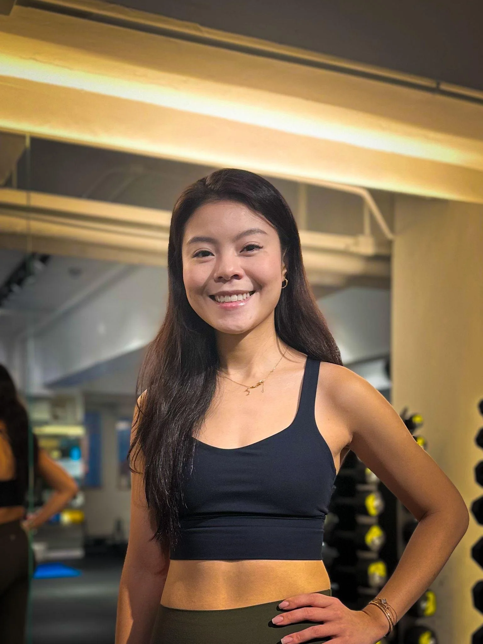 Young woman smiling in gym, wearing black sports bra and olive green leggings.