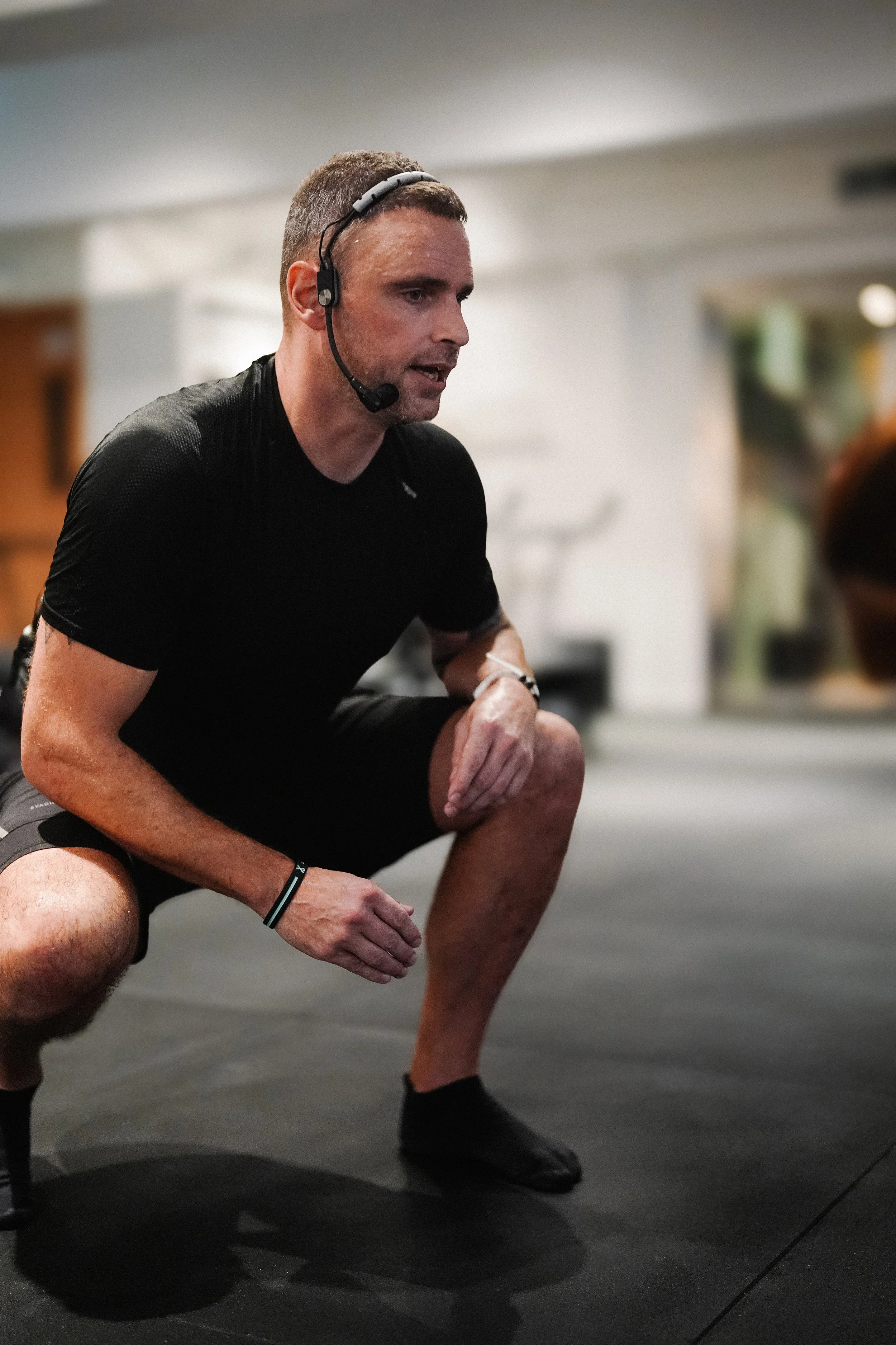 A man wearing a headset crouching on a gym floor, possibly coaching or resting during a workout.