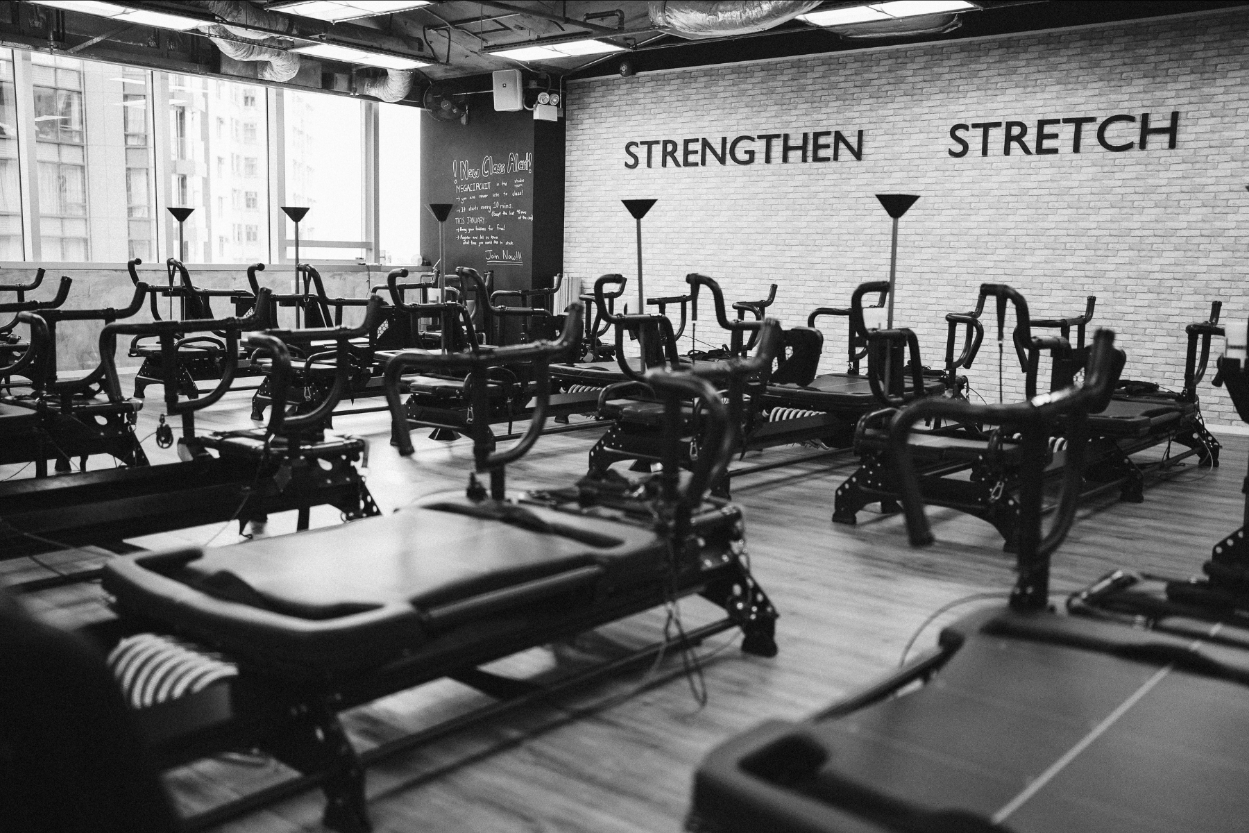Interior of a fitness studio with multiple reformer Pilates machines arranged in rows on a wooden floor, large windows on the left letting in natural light, brick wall with the words 'STRENGTHEN' and 'STRETCH' written on it, and a chalkboard with a motivational message on the back wall.