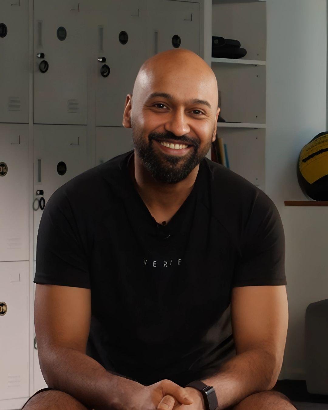 A smiling man with a beard and shaved head sitting inside, wearing a black T-shirt and a smartwatch.