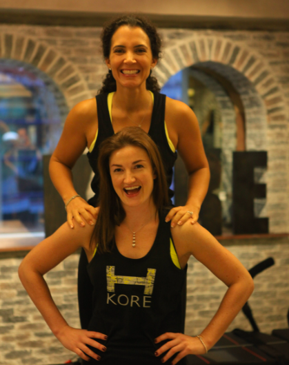 Two women in athletic workout clothes smiling at the camera in a gym with a brick wall background.