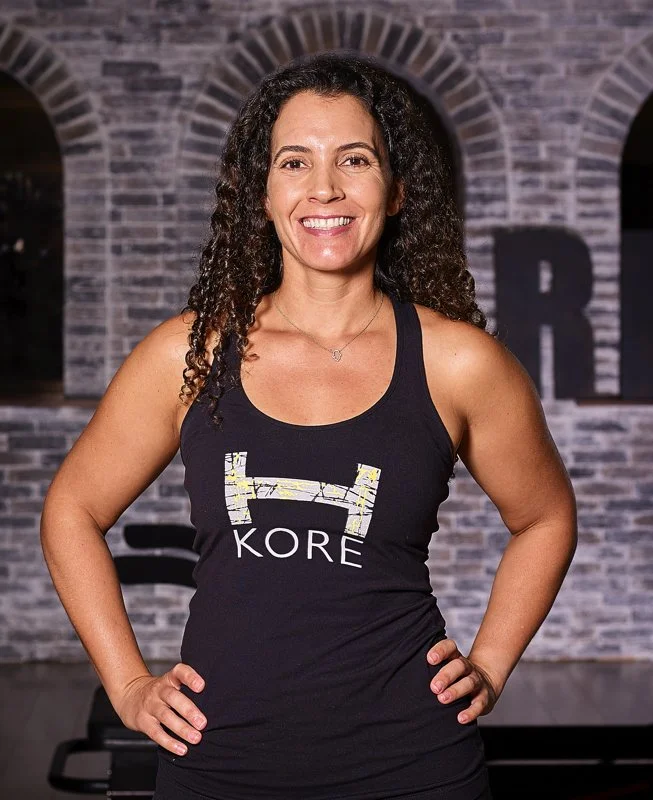 A woman with curly hair smiling, standing with hands on hips in front of a brick wall background.