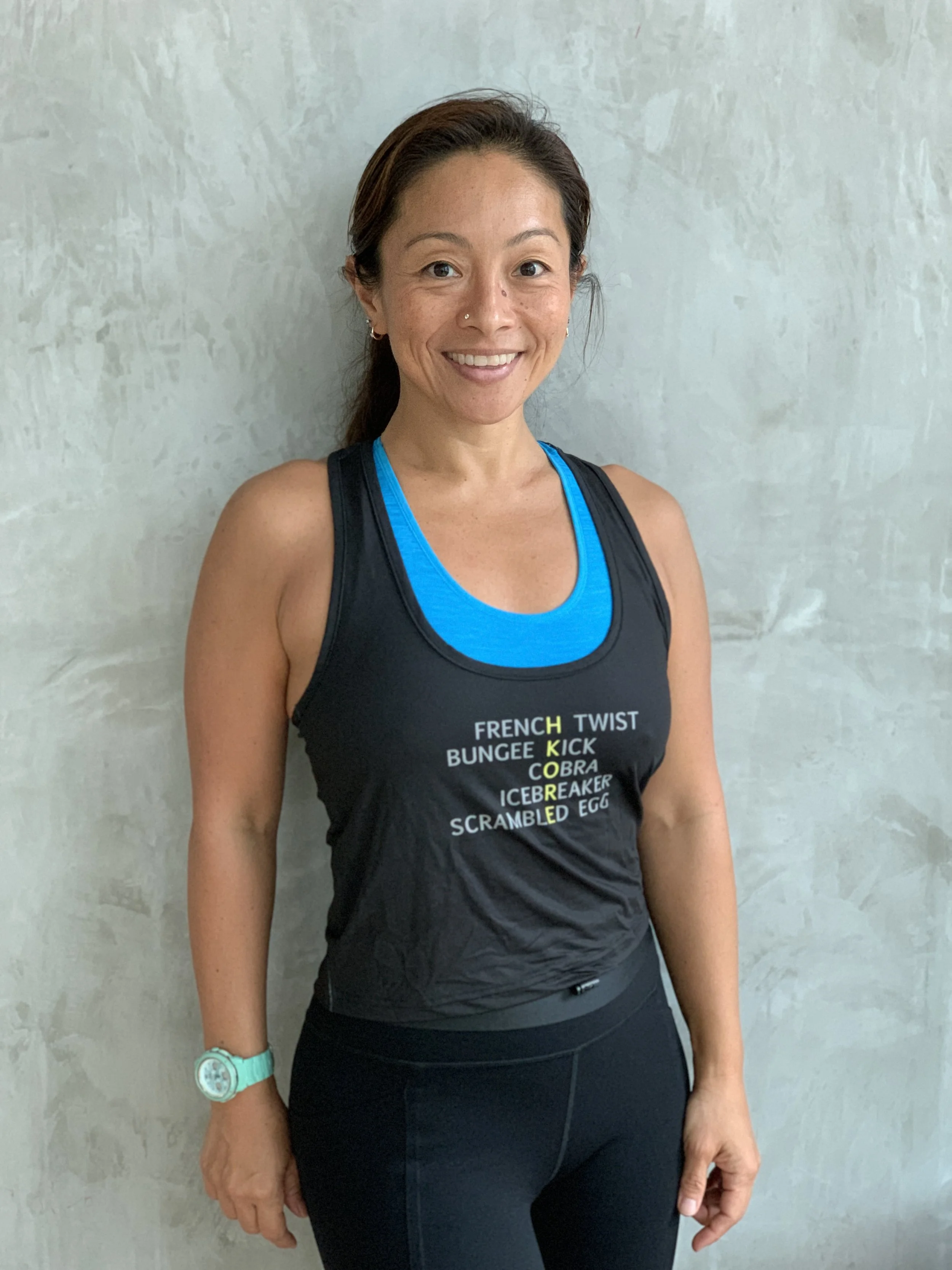 Woman smiling in athletic clothing, wearing a black tank top with workout instructions, and black leggings, standing against a concrete wall.