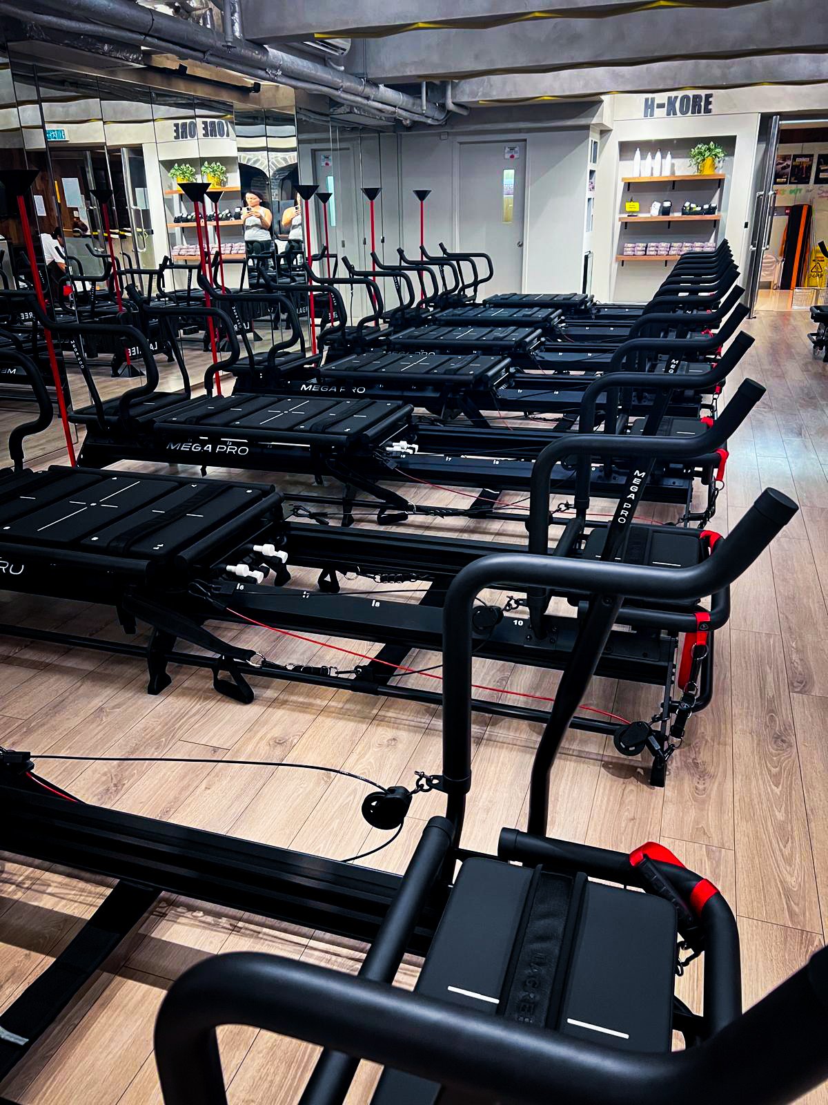 Multiple black exercise machines with handlebars and foot platforms arranged in rows in a fitness studio, with mirrors reflecting the room and a sign that says 'H-KORE' on a shelf in the background.