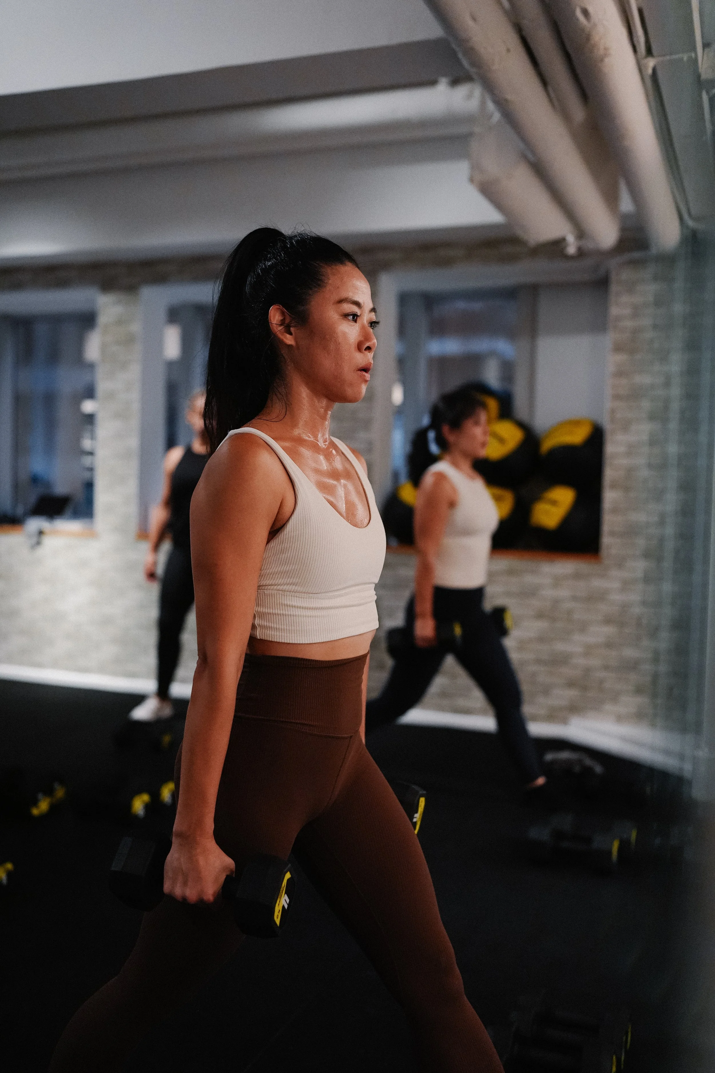 Women exercising in a gym, holding dumbbells, wearing workout clothes, with a serious expression. Two women are visible, one in the foreground and one in the background.