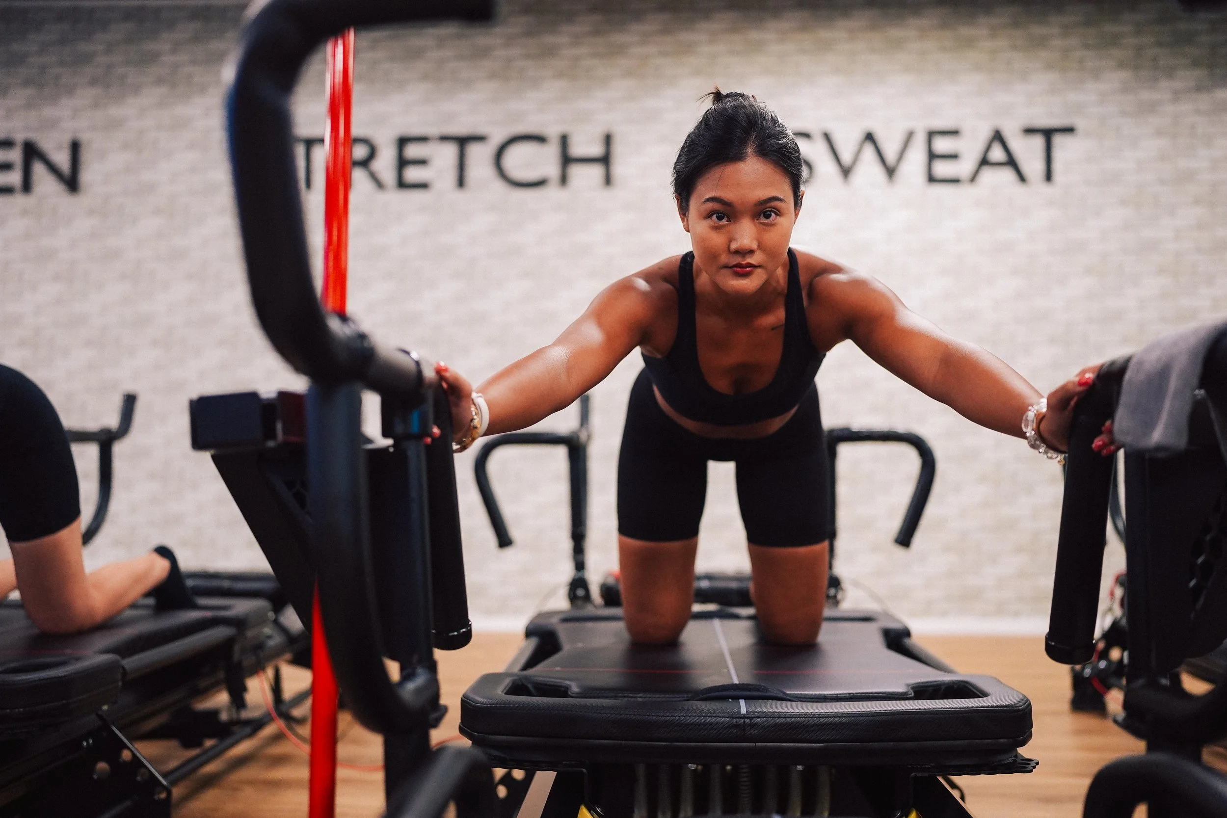 A woman with dark hair in a workout setting, leaning forward on a fitness machine, looking directly at the camera, wearing a black sports bra and black workout shorts.