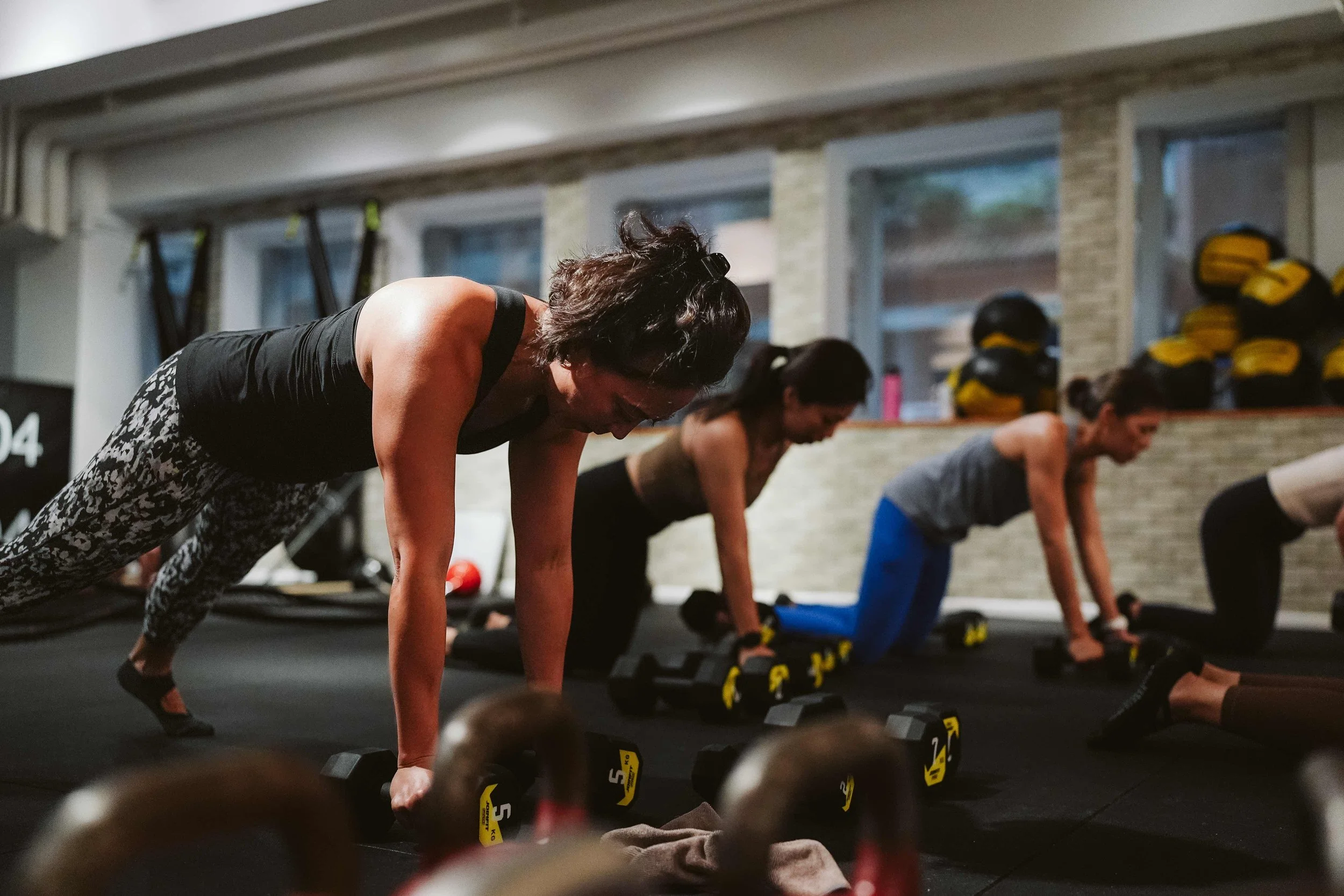Women participating in a group workout class doing planks on mats with dumbbells in a gym.
