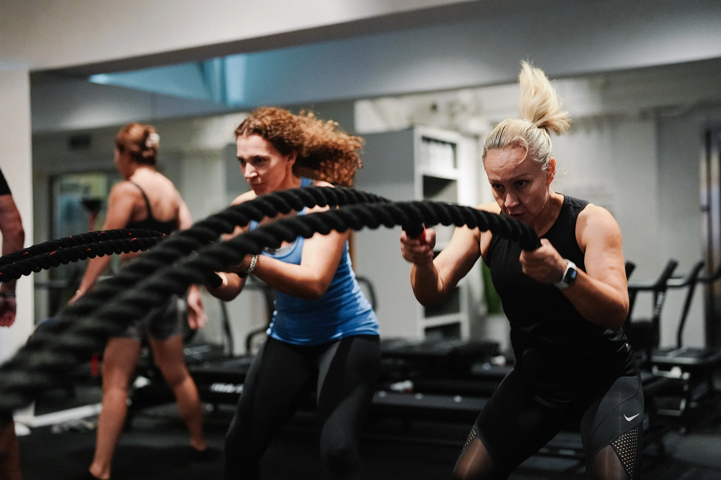 Two women exercising with battle ropes in a gym, both focused and determined during a workout.