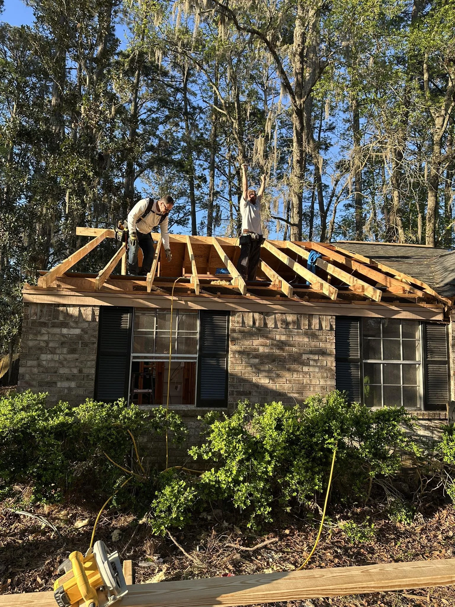 Two construction workers are working on a house roof, installing wooden rafters with a backdrop of trees and blue sky.