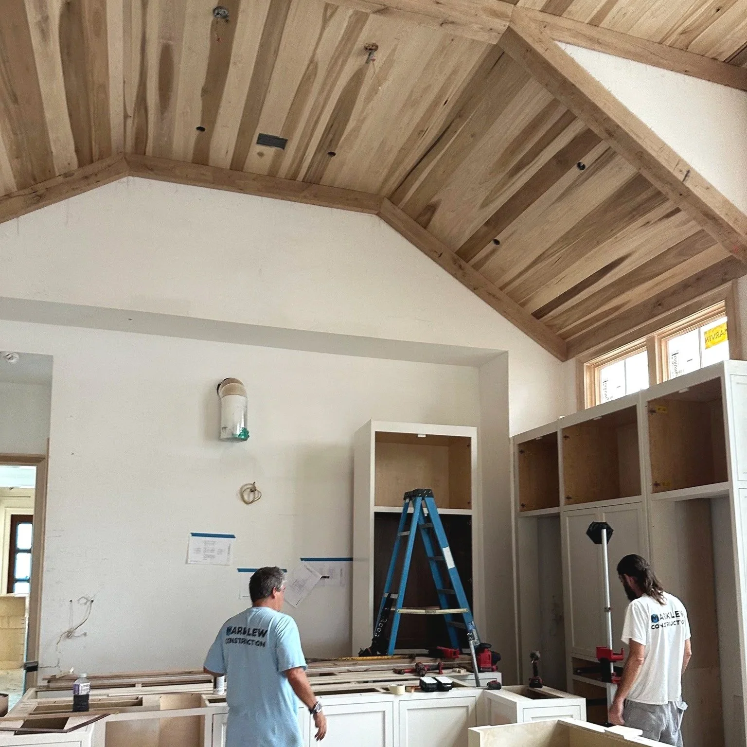 Construction workers inside a room with unfinished cabinetry and a vaulted wooden ceiling with white walls.