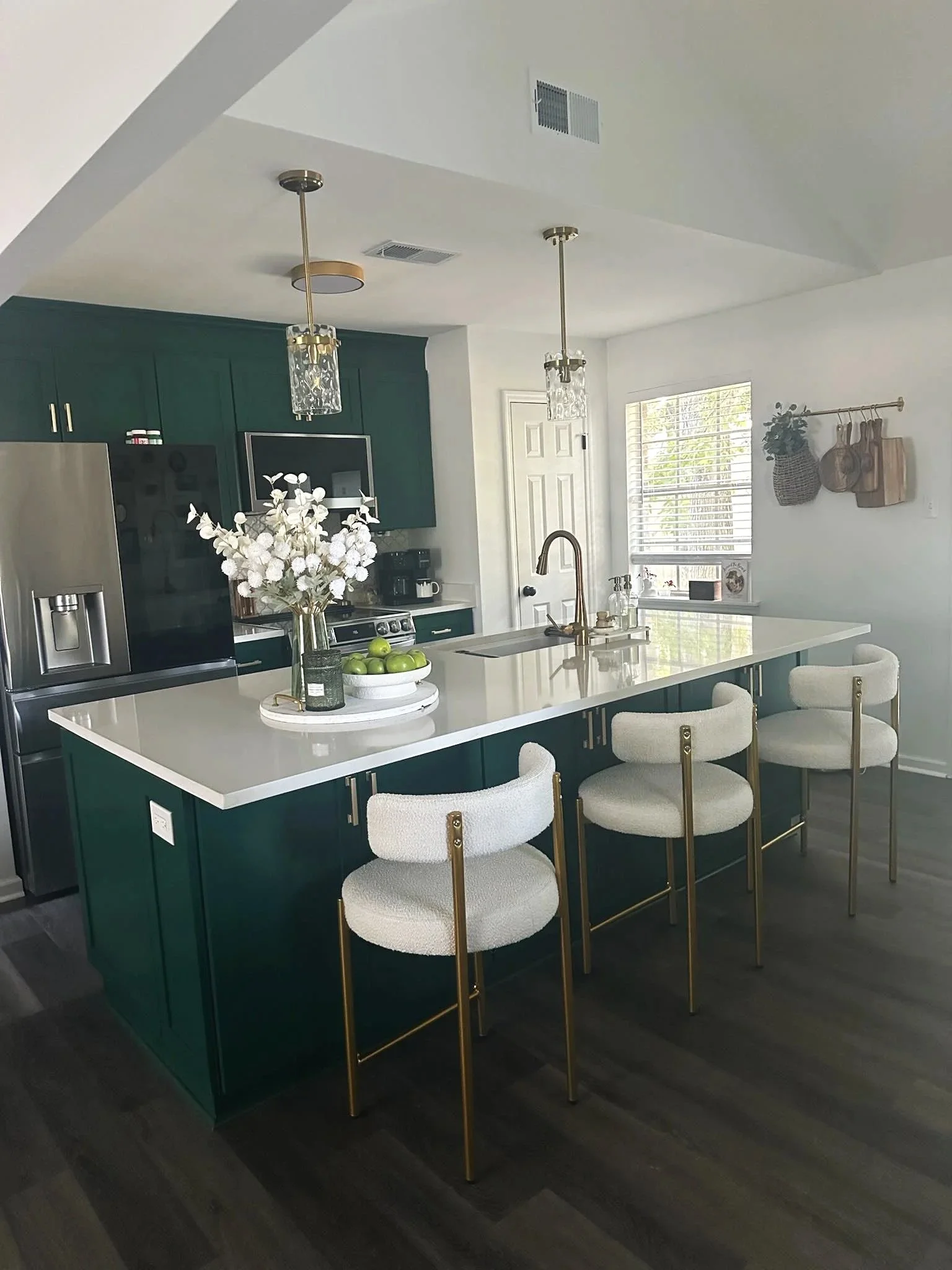 Modern kitchen with a green island, white countertop, four white bar stools with gold accents, dark wood flooring, and hanging pendant lights.