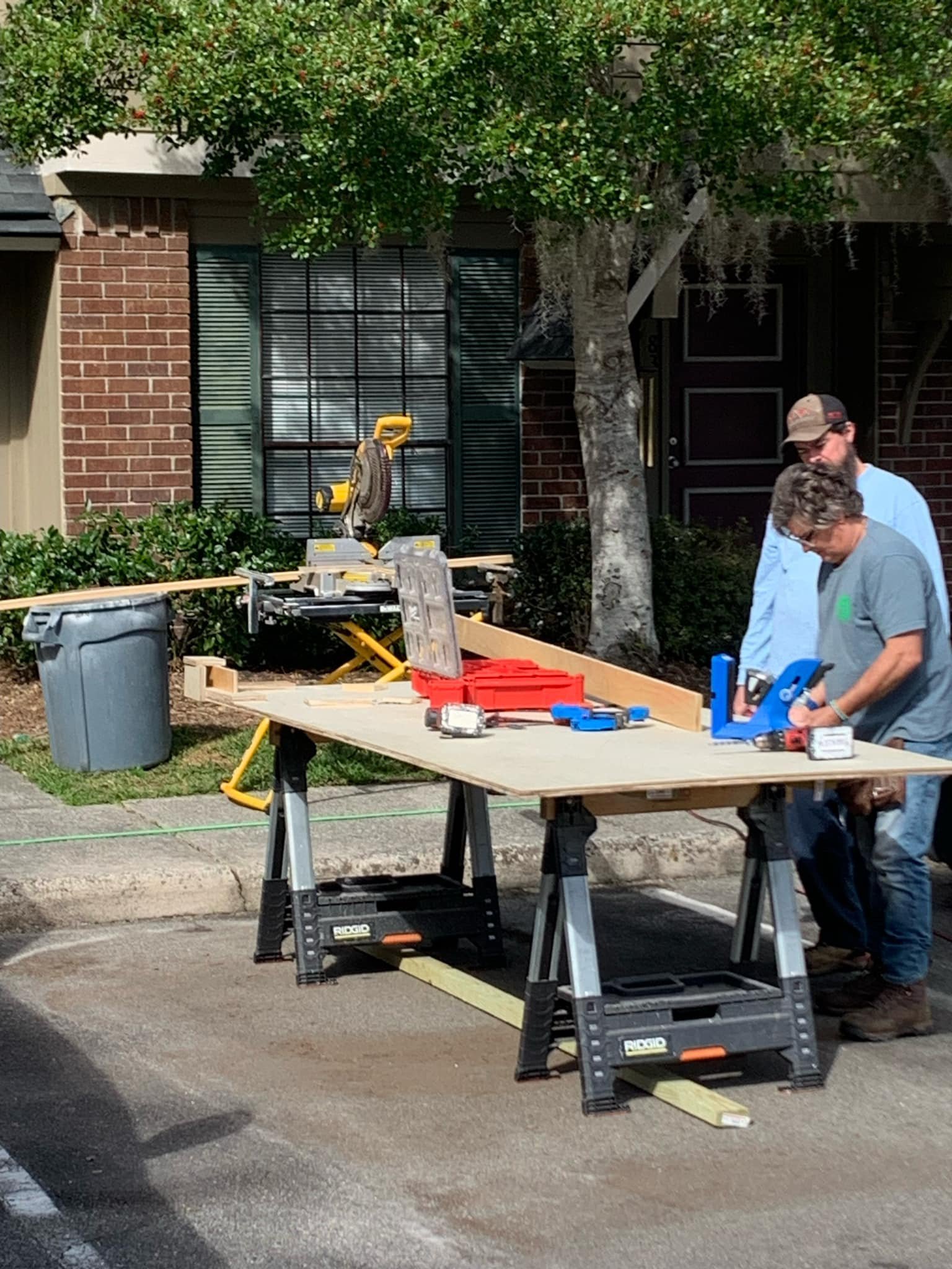 Two people are working on a home improvement project outdoors, using power tools on a wooden workbench set up on a parking lot, with building and trees in the background.