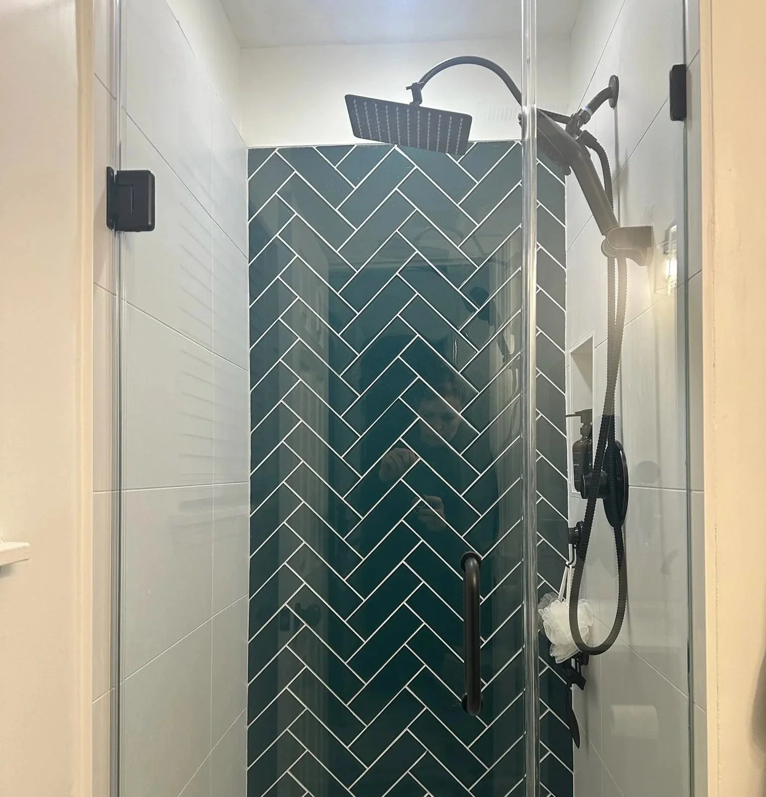 Inside a modern shower with a glass door, blue herringbone tile pattern on the back wall, a black showerhead, and a handheld shower wand with a white shower sponge hanging from it.