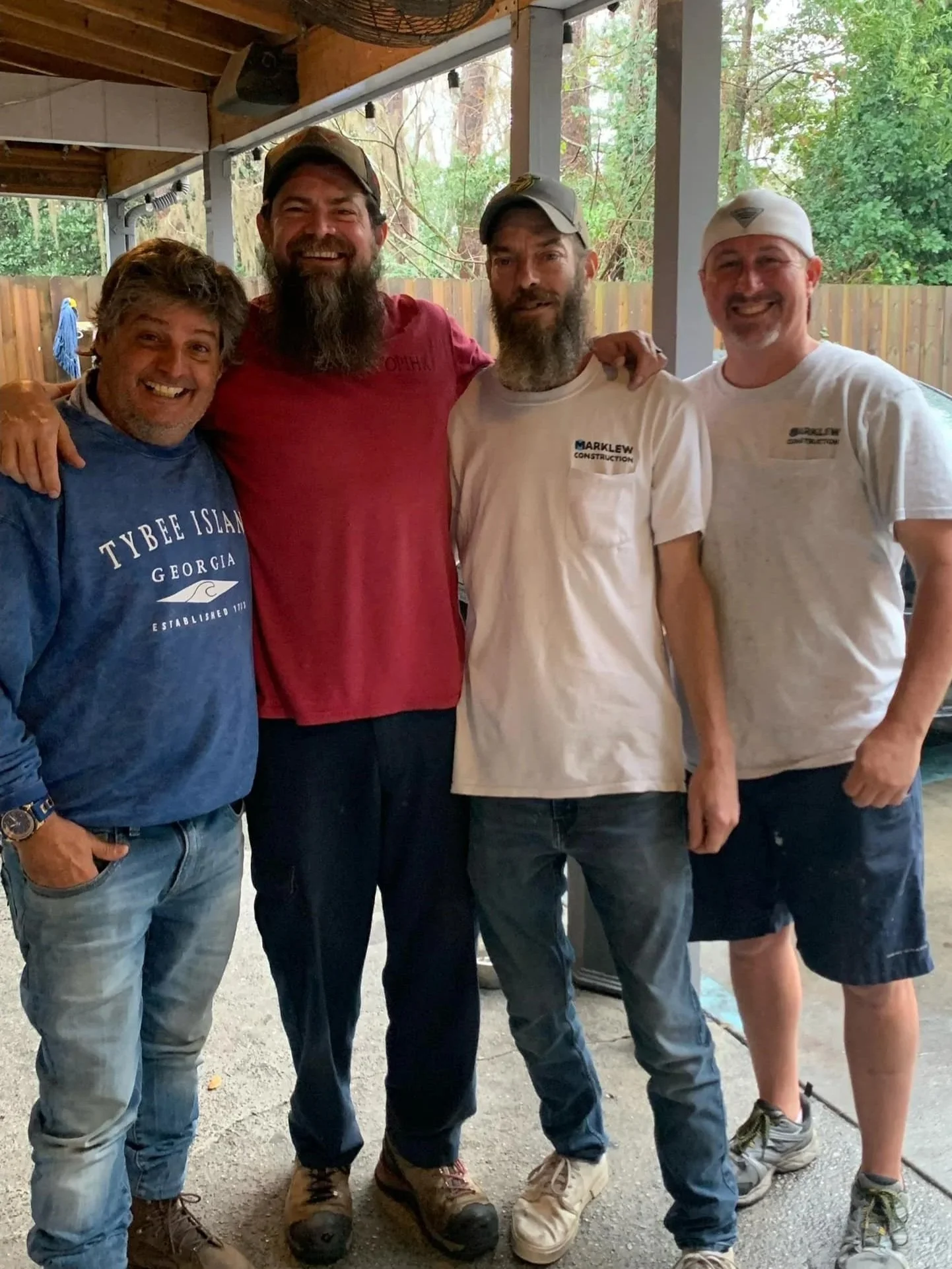 Four men standing together inside a wooden structure, smiling at the camera. One man is wearing a blue hoodie that says 'Tybee Island Georgia,' another in a red T-shirt, and two in white T-shirts with 'Marklew Construction' logos. They are outdoors, with trees and a wooden fence visible in the background.
