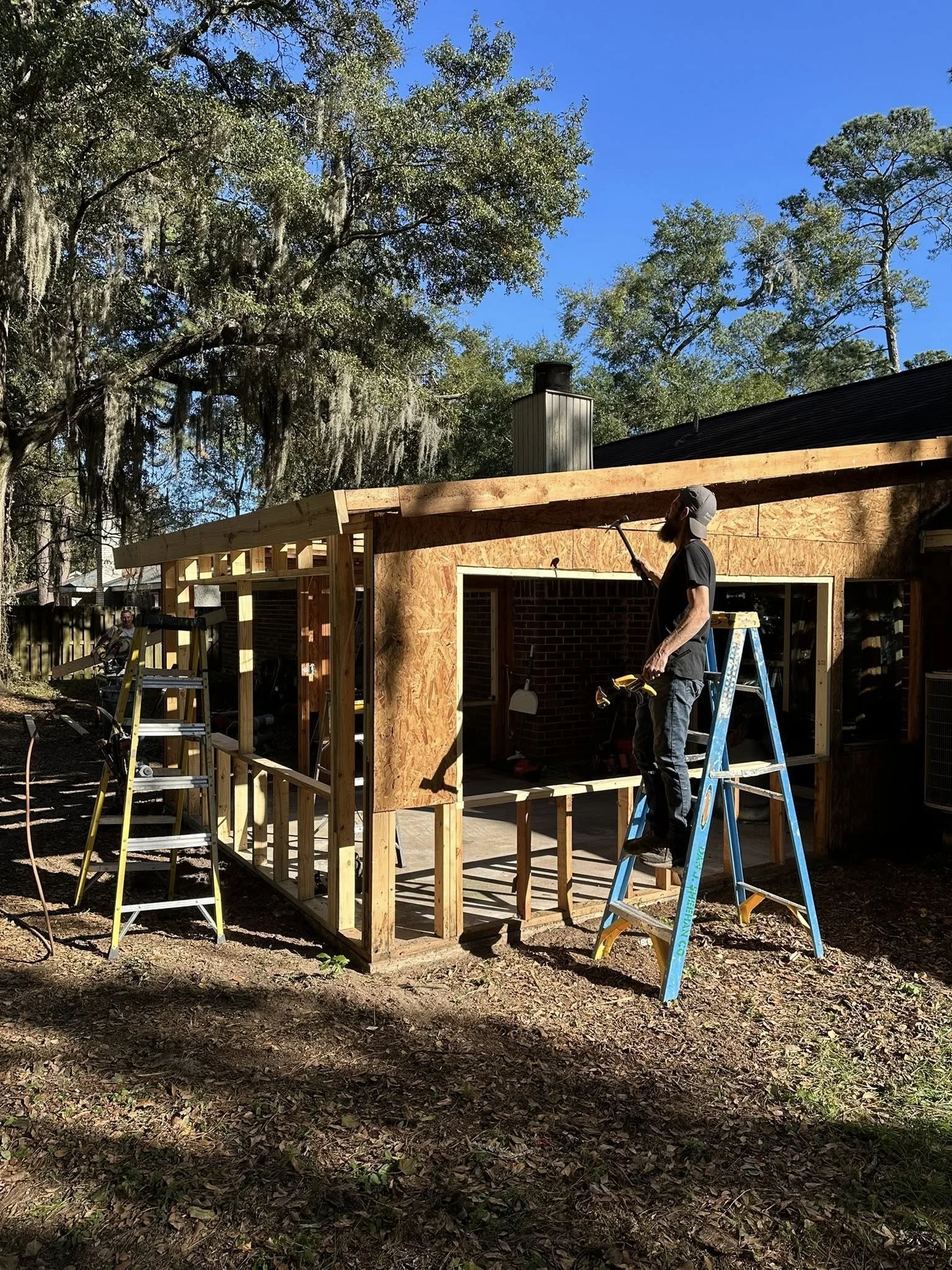 Person standing on a ladder, working on a wooden porch extension attached to a house, with construction tools nearby, in a backyard with trees.