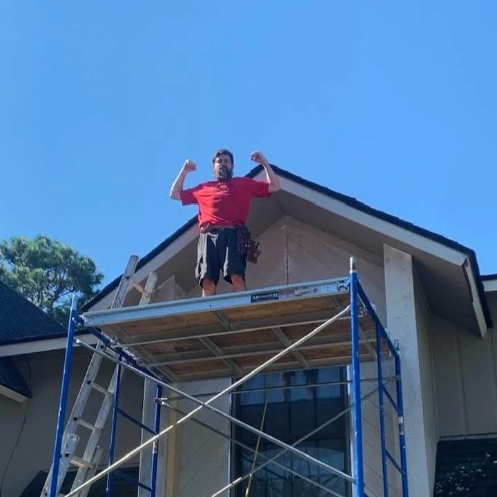 A man standing on a scaffold in front of a house, flexing his biceps.
