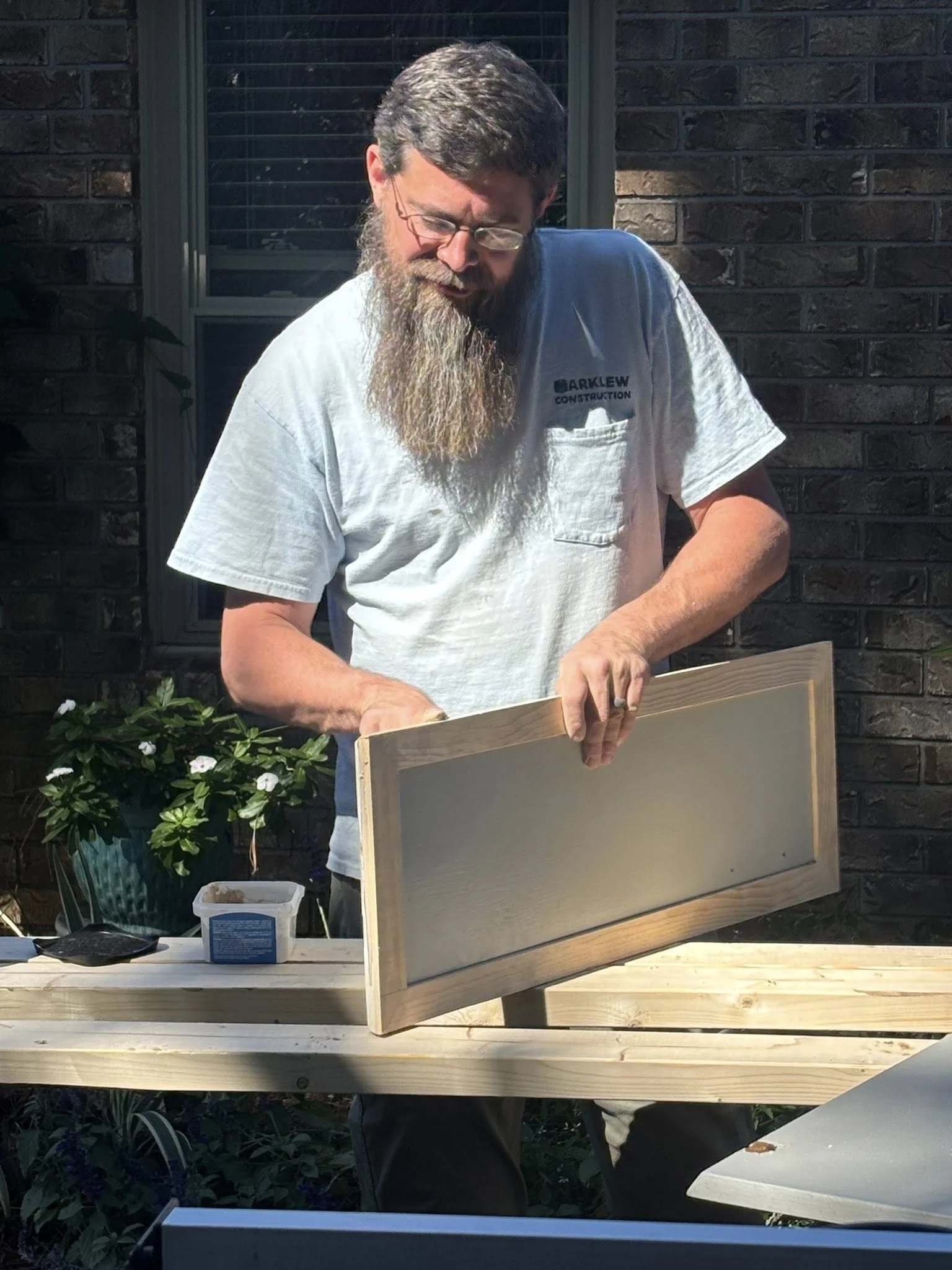 A man with a beard and glasses arranging a wooden panel outdoors with a brick house and garden in the background.